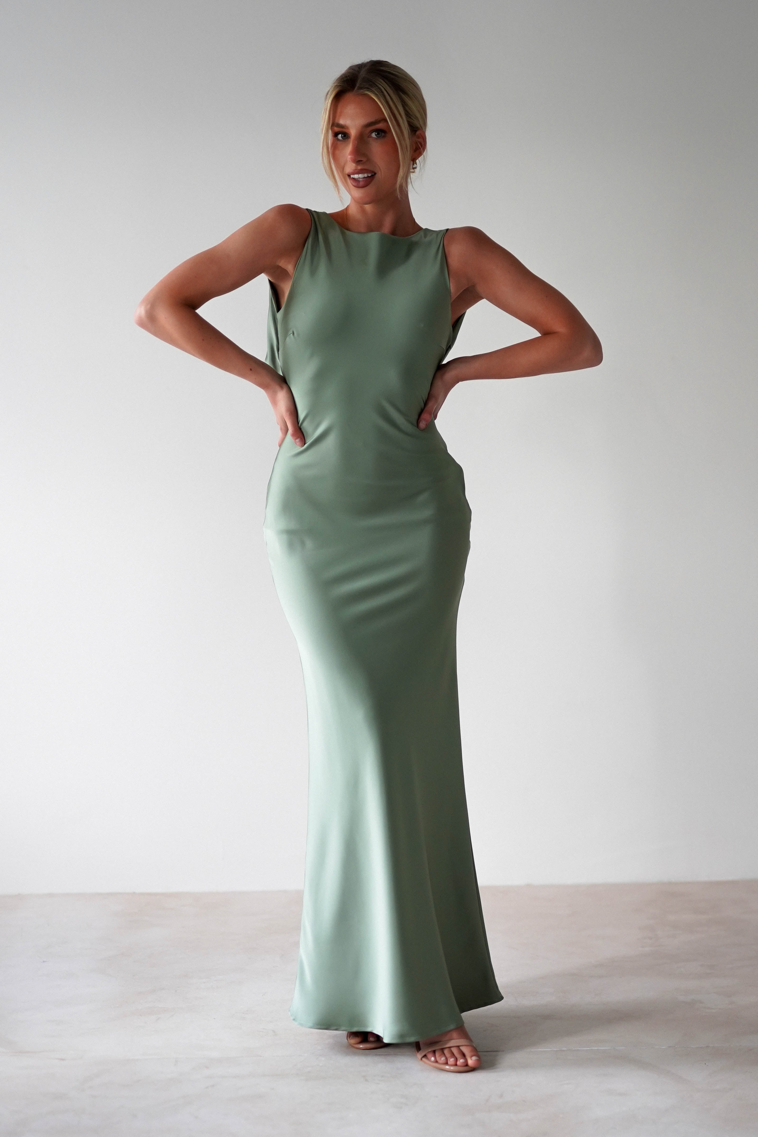 A woman stands indoors wearing a sleeveless, floor-length light green satin maxi dress with her hands on her hips, looking confidently at the camera. The stylish gown is perfect as a wedding guest dress. The background is plain and light-colored.