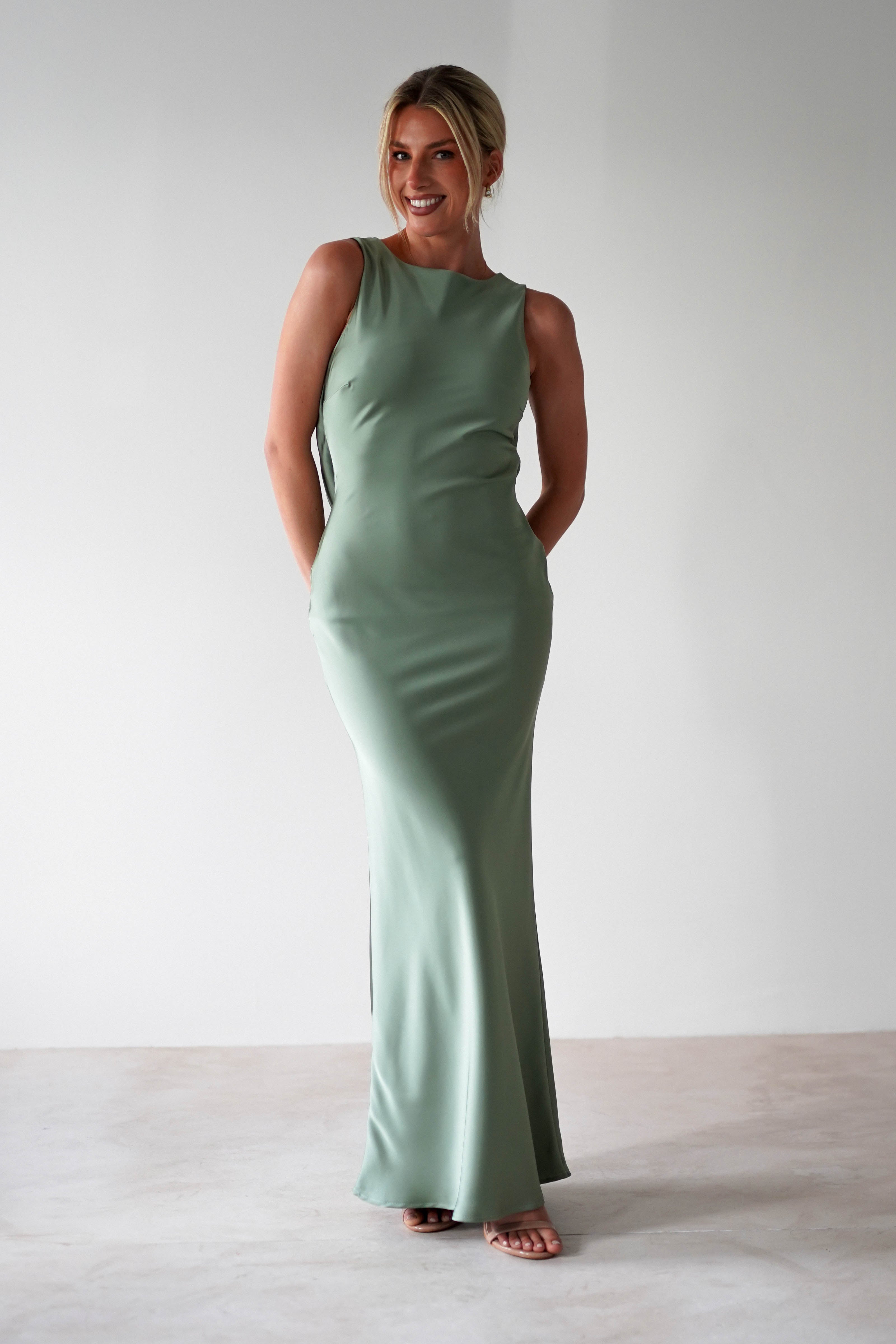 A woman stands smiling in a sleeveless, light green satin maxi dress with her hands in the pockets, against a plain white background.