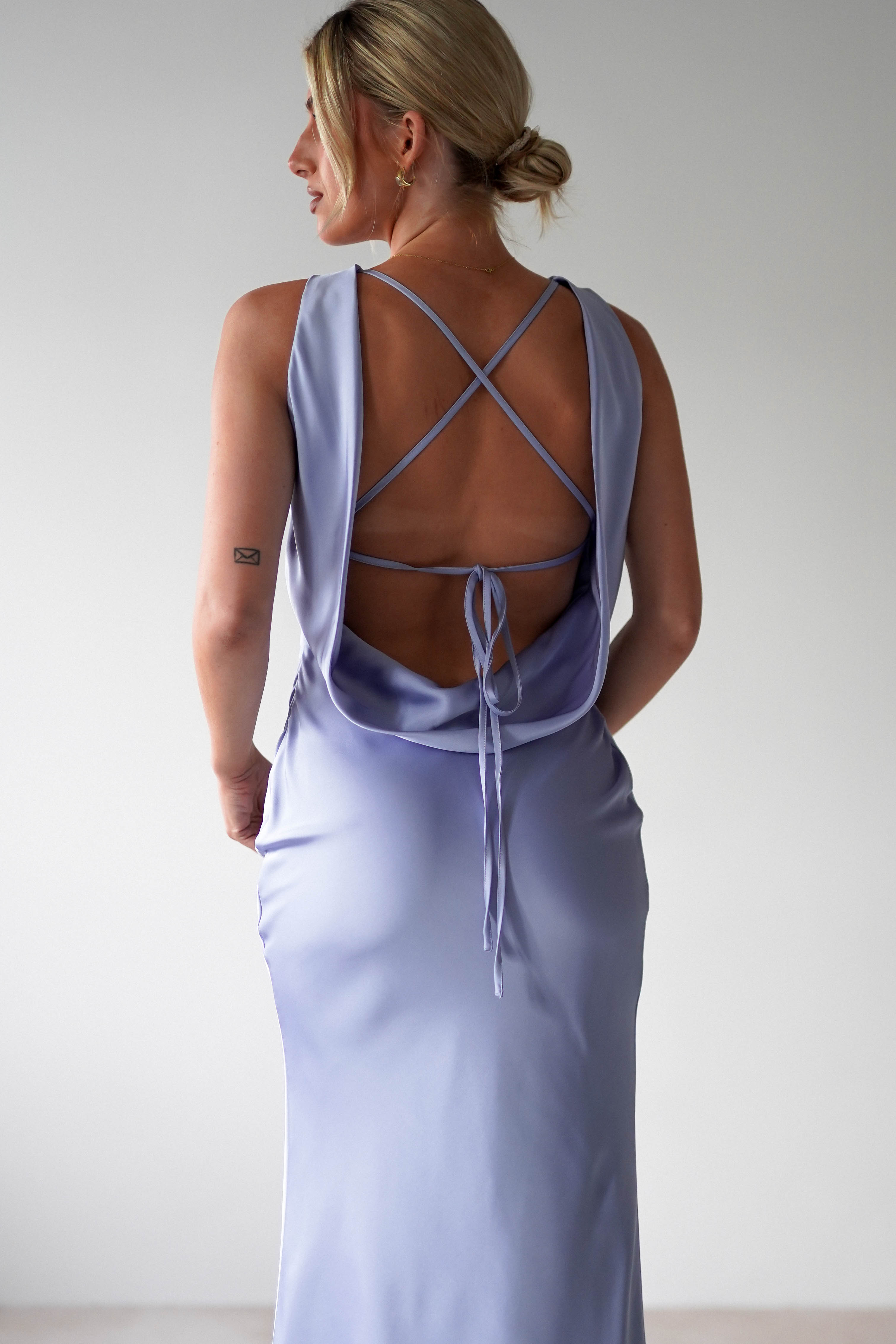 A person with blonde hair stands with their back to the camera, draped in a light blue satin maxi dress. The elegant gown features a low, open back with crisscross straps and a delicate tie. A small tattoo graces their arm against the plain, light-colored backdrop, evoking bridesmaid charm.