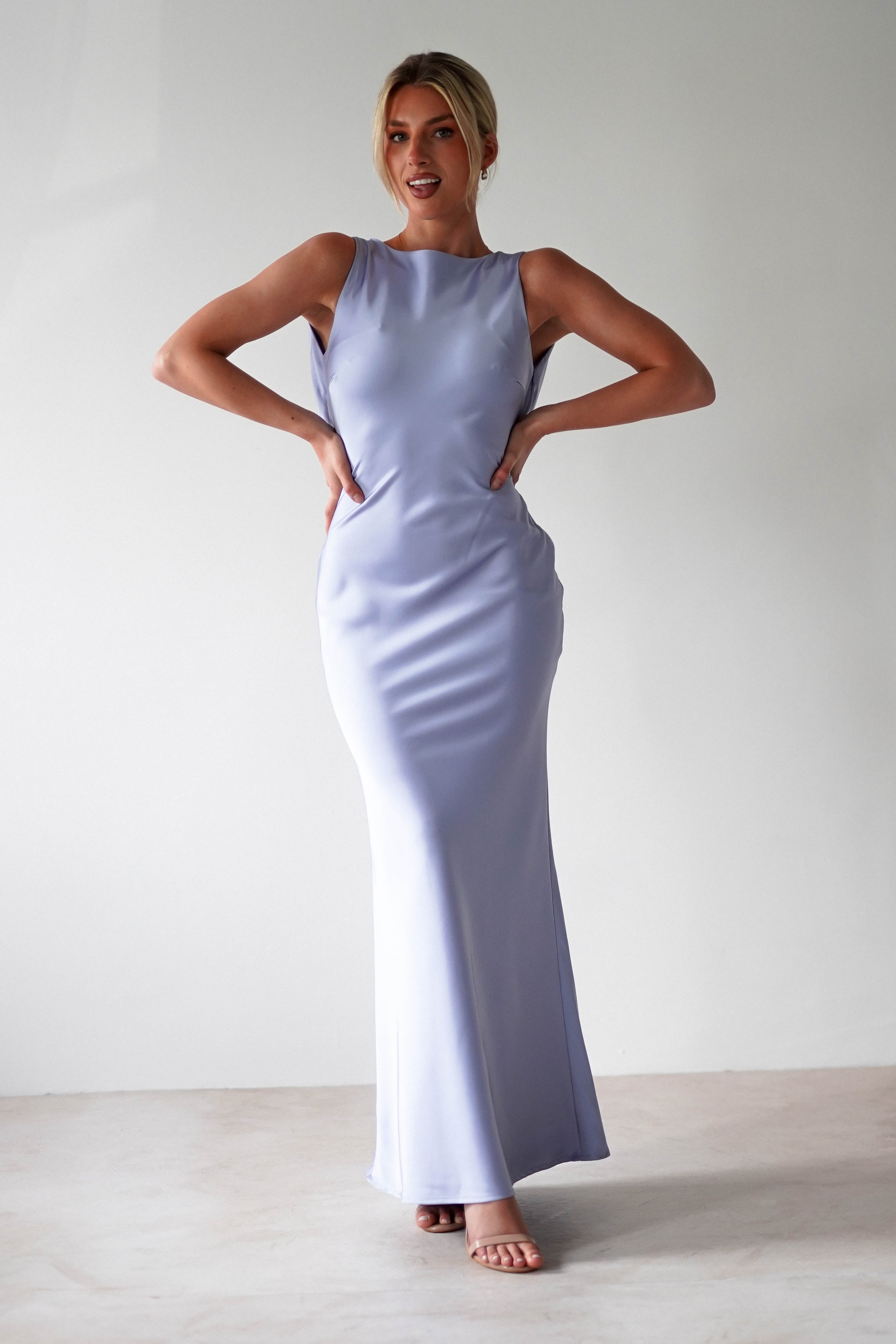 A person stands confidently in a sleek, light purple satin maxi dress with an open back, showcasing a fitted silhouette and high neckline. Their hands rest on their hips, complemented by open-toed heels. The background remains plain and neutral.