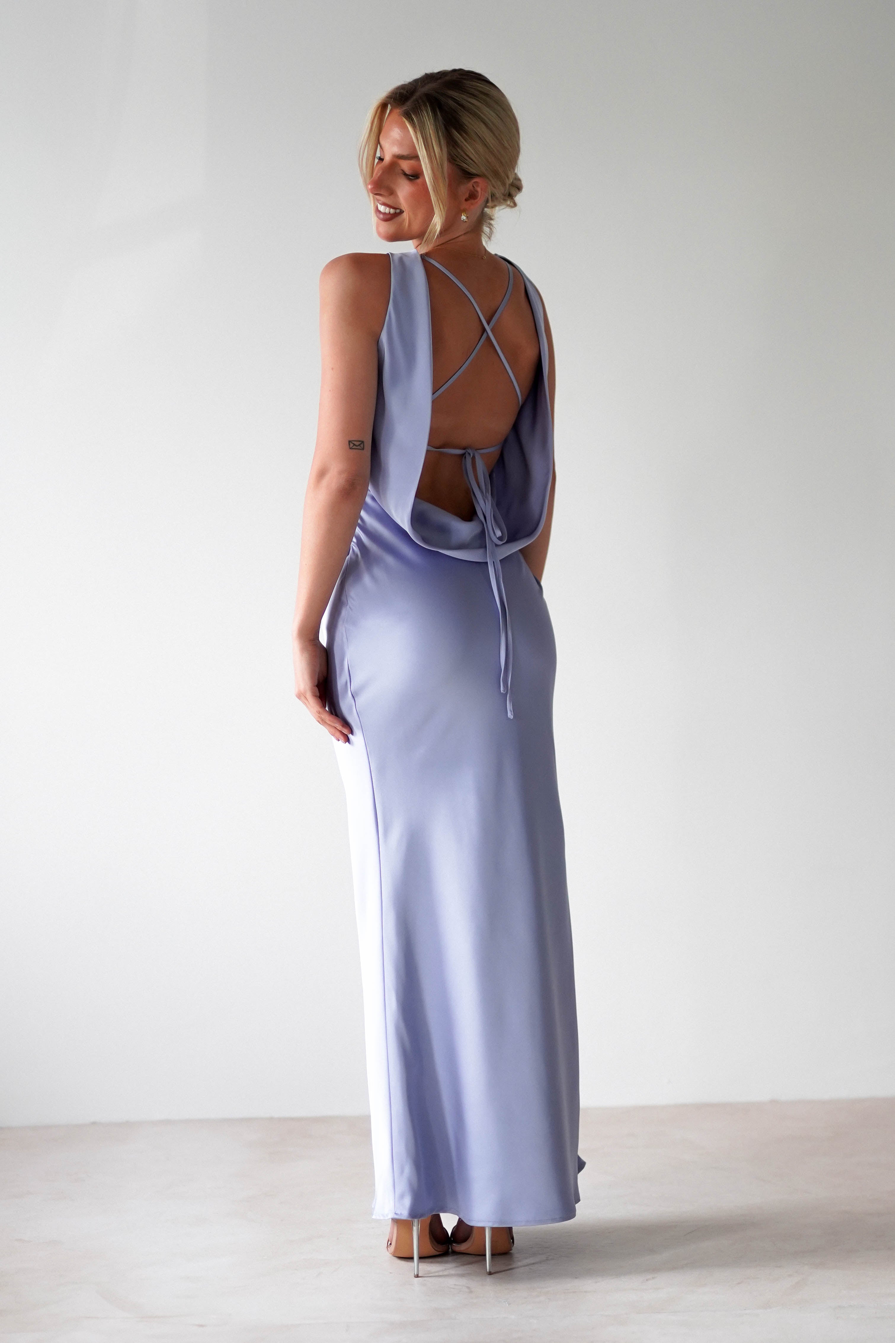A woman in a light blue satin maxi dress with an open back stands on a light floor. She is smiling and looking over her shoulder, showcasing the crisscross back design and tied detail of the dress, perfect for bridesmaids. Her hair is styled up.