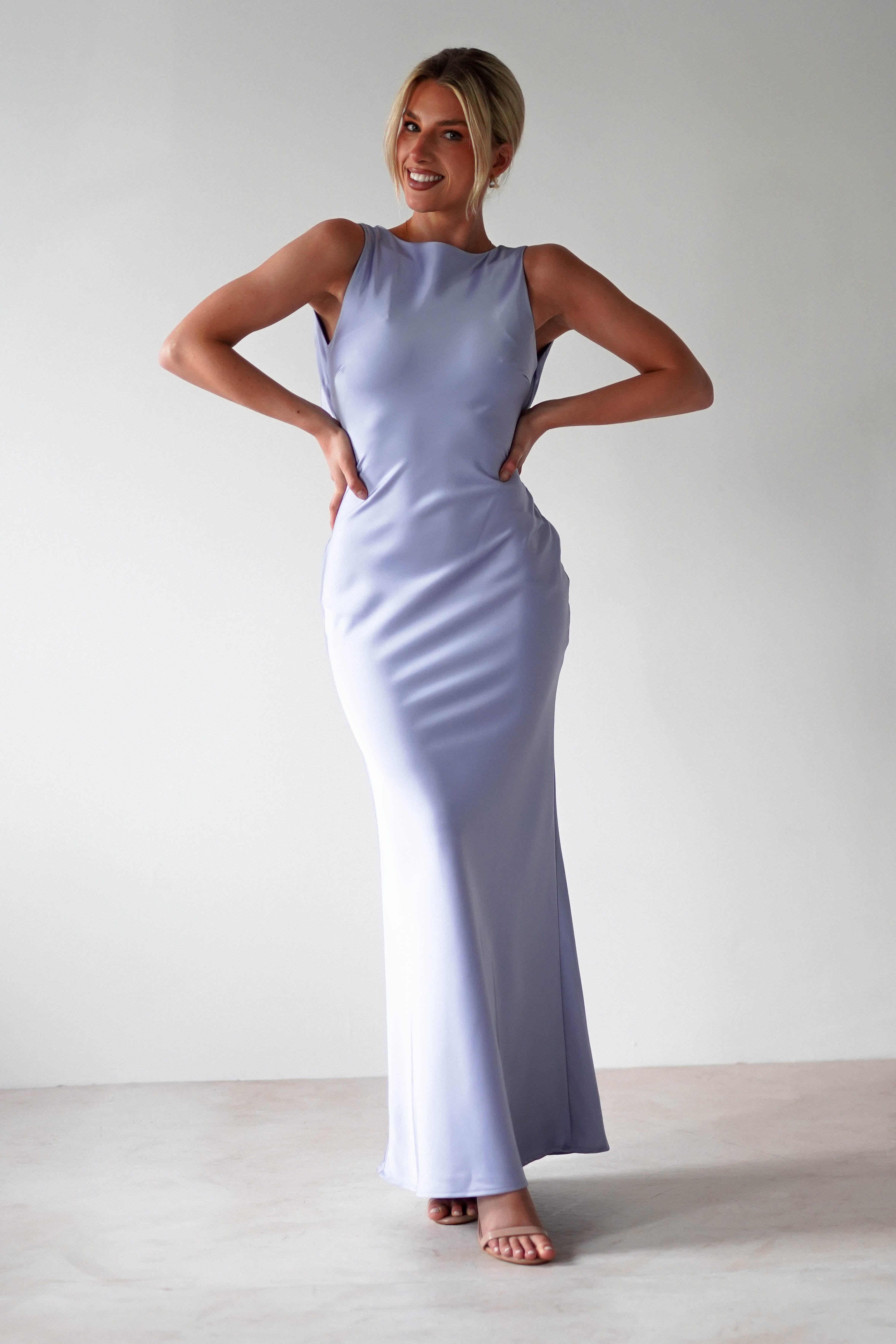 A person stands smiling, wearing a sleeveless, satin maxi dress in light blue. The gown features an elegant open back and hugs the form beautifully. Their hair is styled up, and they pose confidently with hands on hips against a plain, light-colored background.