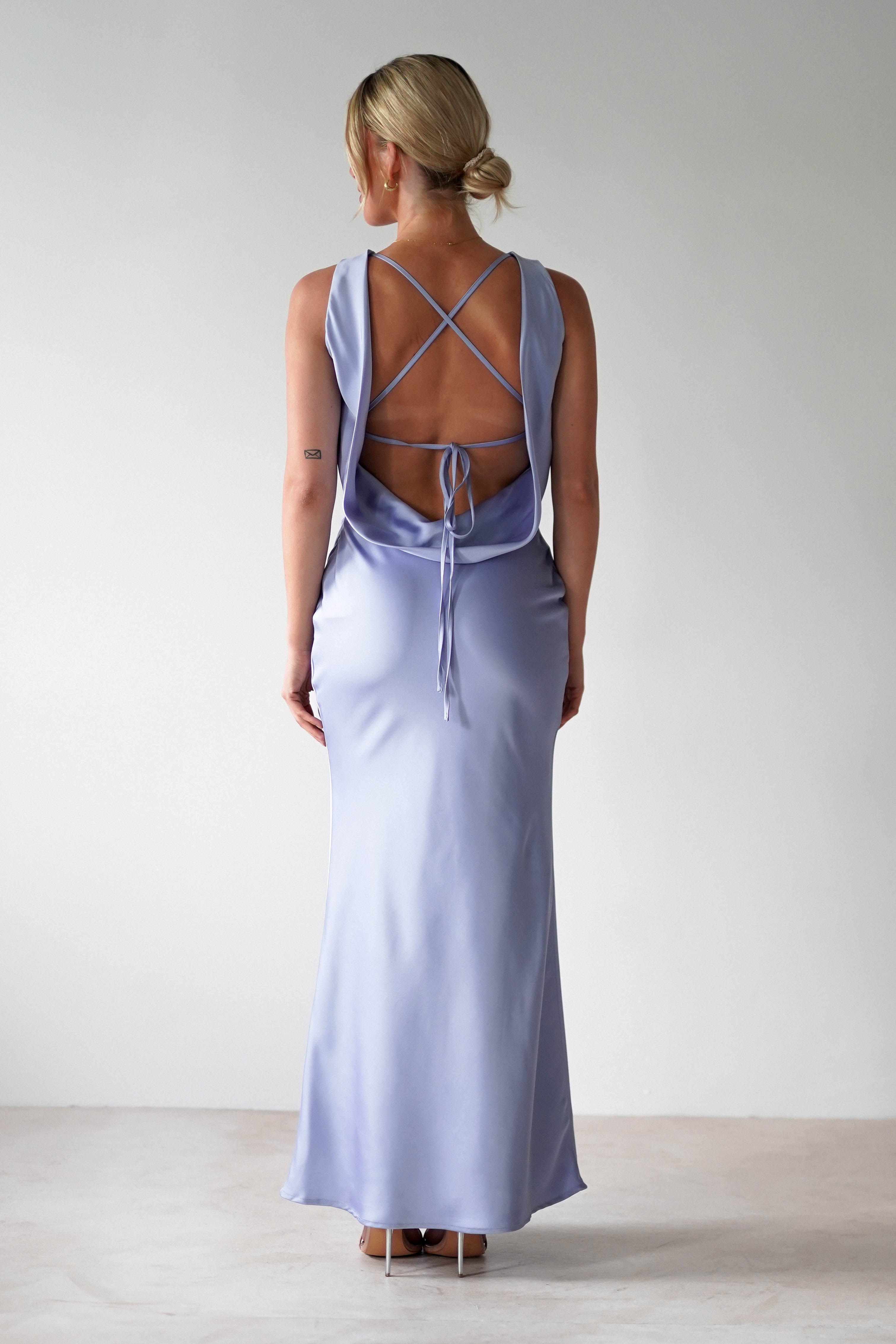 Rear view of a person in a light purple satin maxi dress with an open back and crisscross straps. They have blonde hair styled in a low bun and are wearing high heels, perfect for bridesmaids. The setting is a plain, light-colored room.