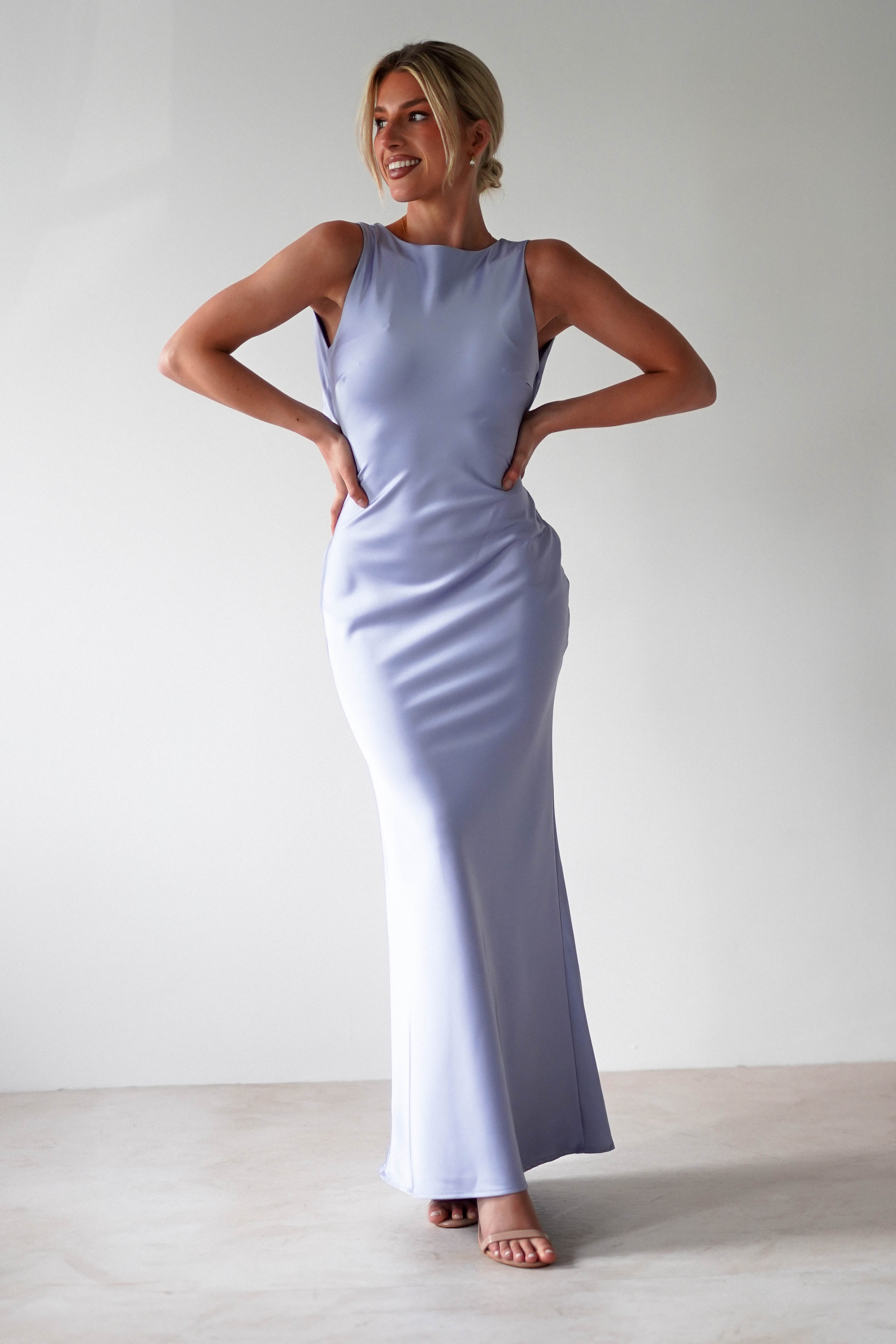 A person wearing a sleeveless, floor-length satin maxi dress in lavender stands with their hands on their hips, smiling. The elegant gown has a subtle sheen and an open back. They have short, light-colored hair and pose against a plain white background.