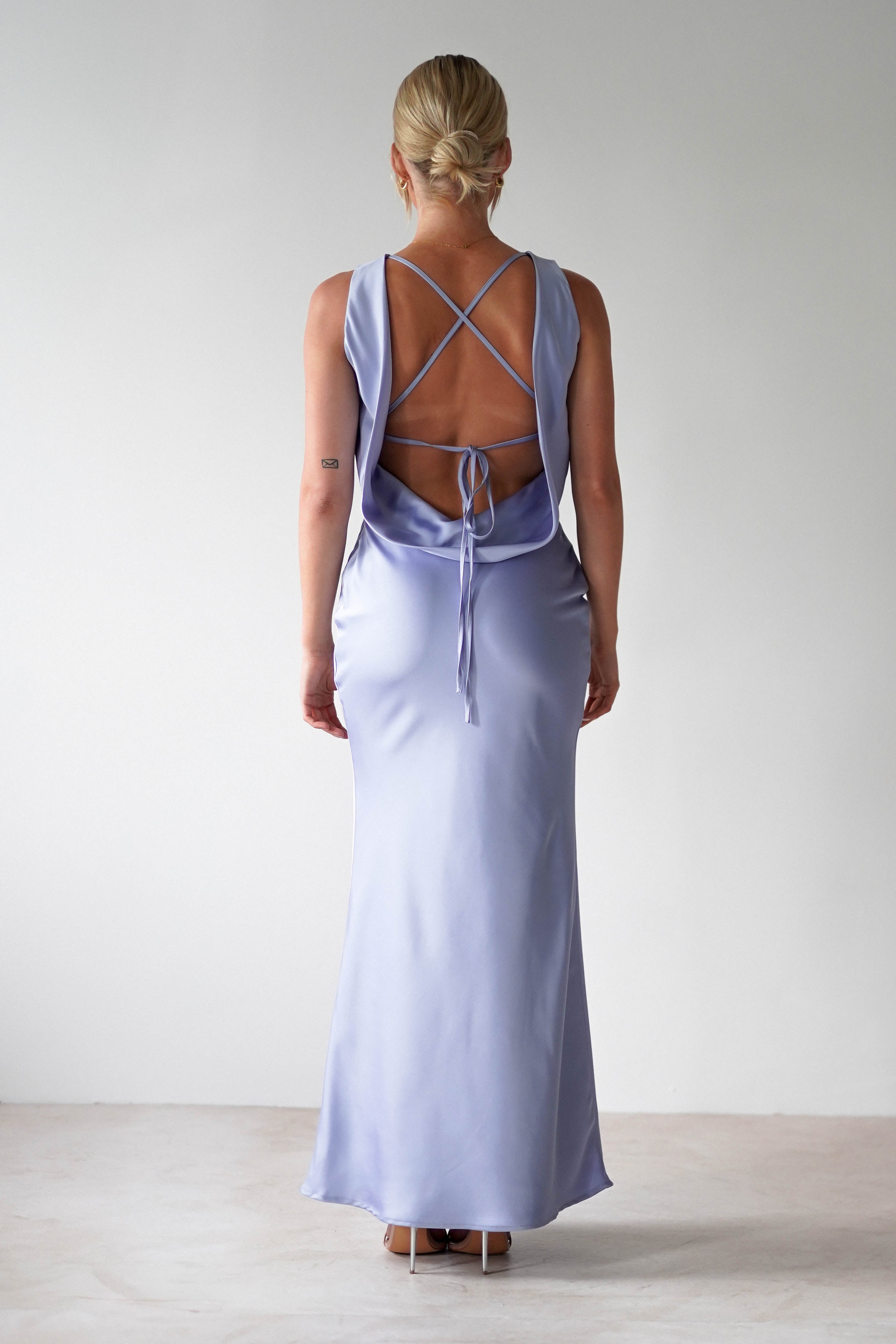 A woman stands facing away, dressed in an elegant satin maxi dress in light lavender with crisscross straps and an open tie back. Her hair is styled in an updo, and she wears high heels. The simple elegance of the gown makes it perfect for bridesmaids against the plain, light-colored wall backdrop.