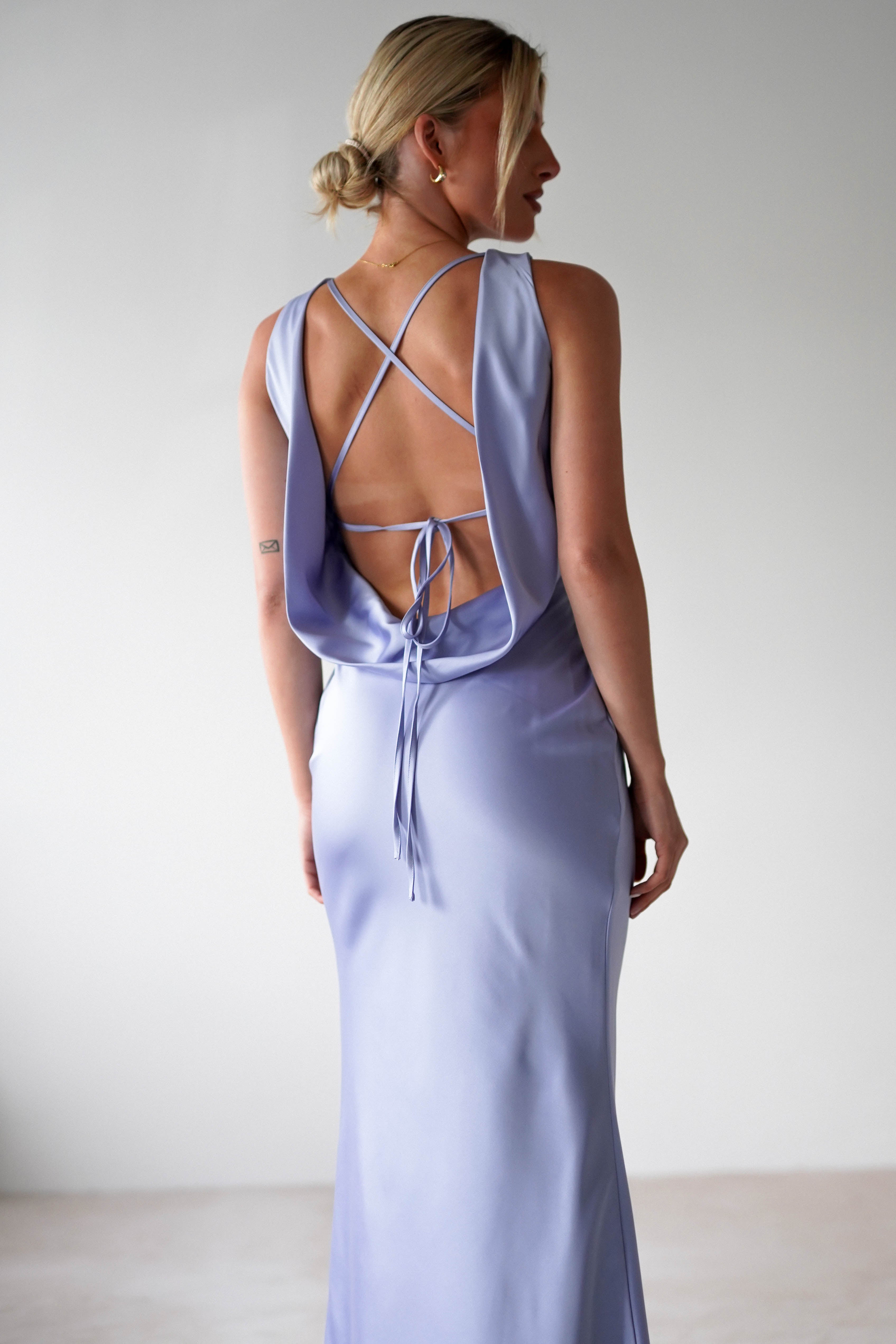 A woman stands with her back to the camera in a pale lavender satin maxi dress, featuring thin crisscross straps and tie detailing. Her blonde hair is styled in a low bun, perfectly complementing the minimalist setting with its neutral background, ideal for bridesmaids seeking elegance.