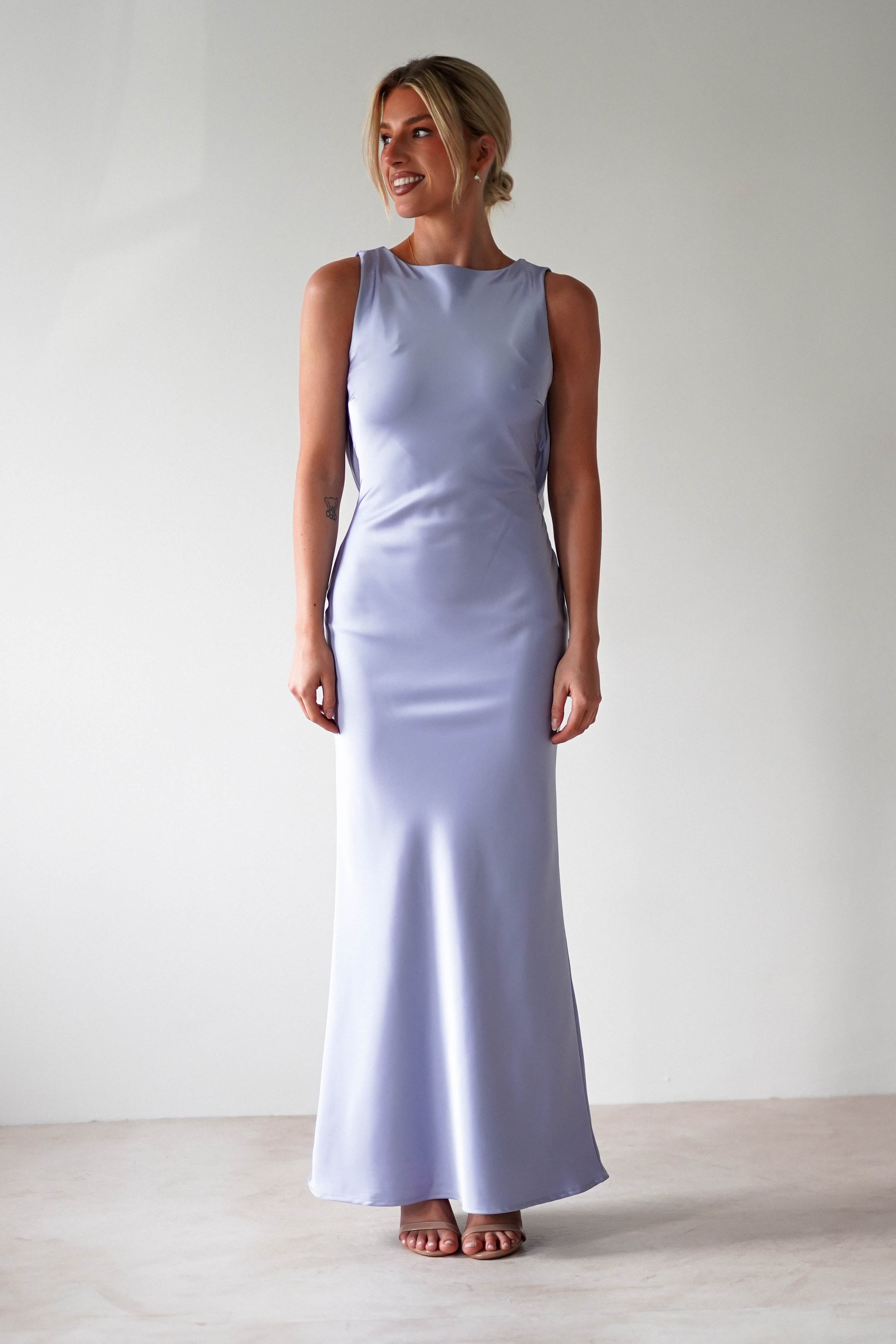 The woman is elegantly dressed in a sleeveless, floor-length satin maxi dress with an open back, perfect for bridesmaids. Shes standing against a plain white background, smiling with her hair styled in an updo and wearing chic open-toed sandals.