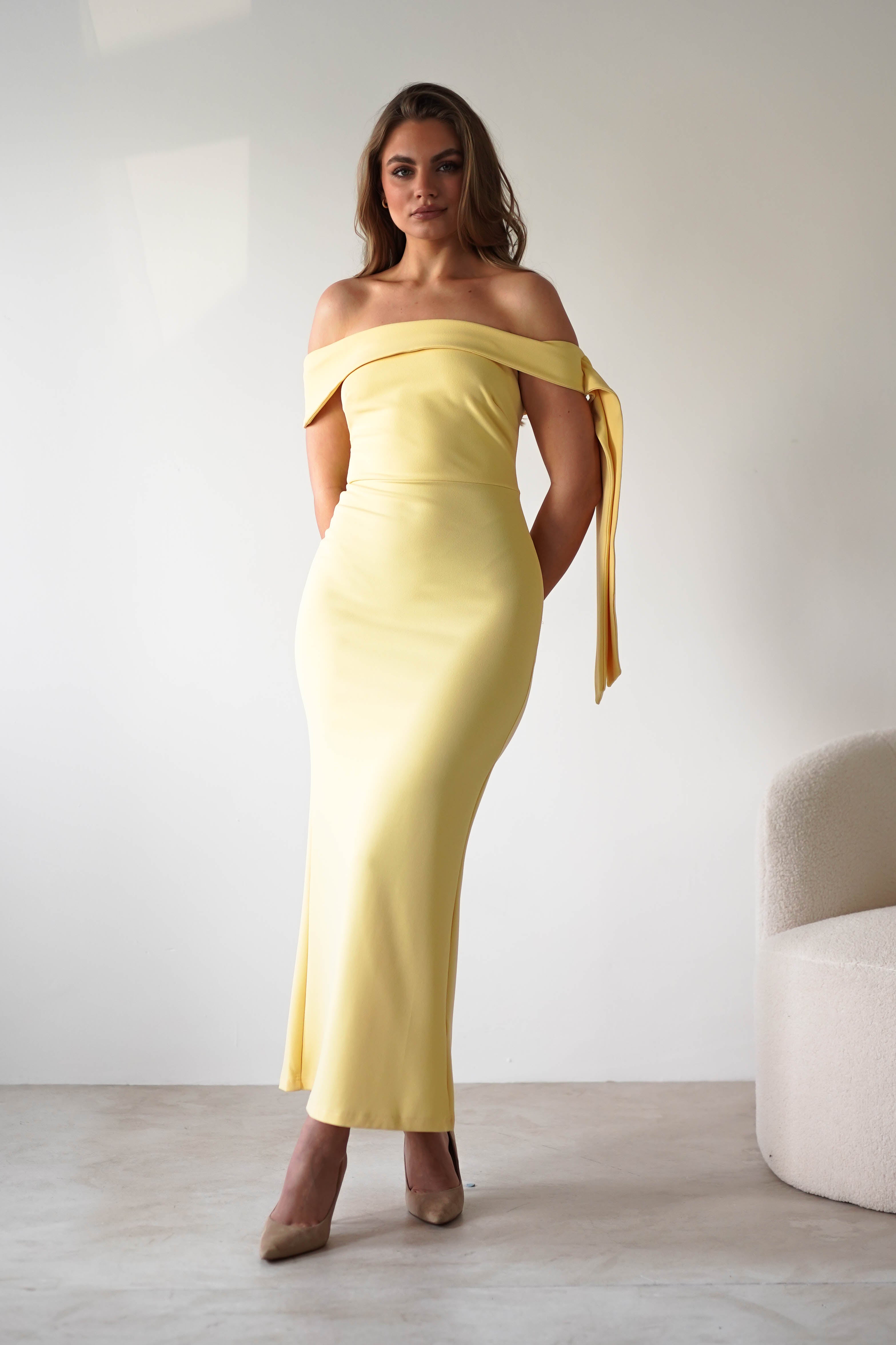 A woman stands indoors against a white wall, wearing a long, off-the-shoulder dress with an A-line skirt and a bow detail on one sleeve. With light brown hair and beige heels, she looks perfect for a wedding guest. A cream-colored chair is nearby.