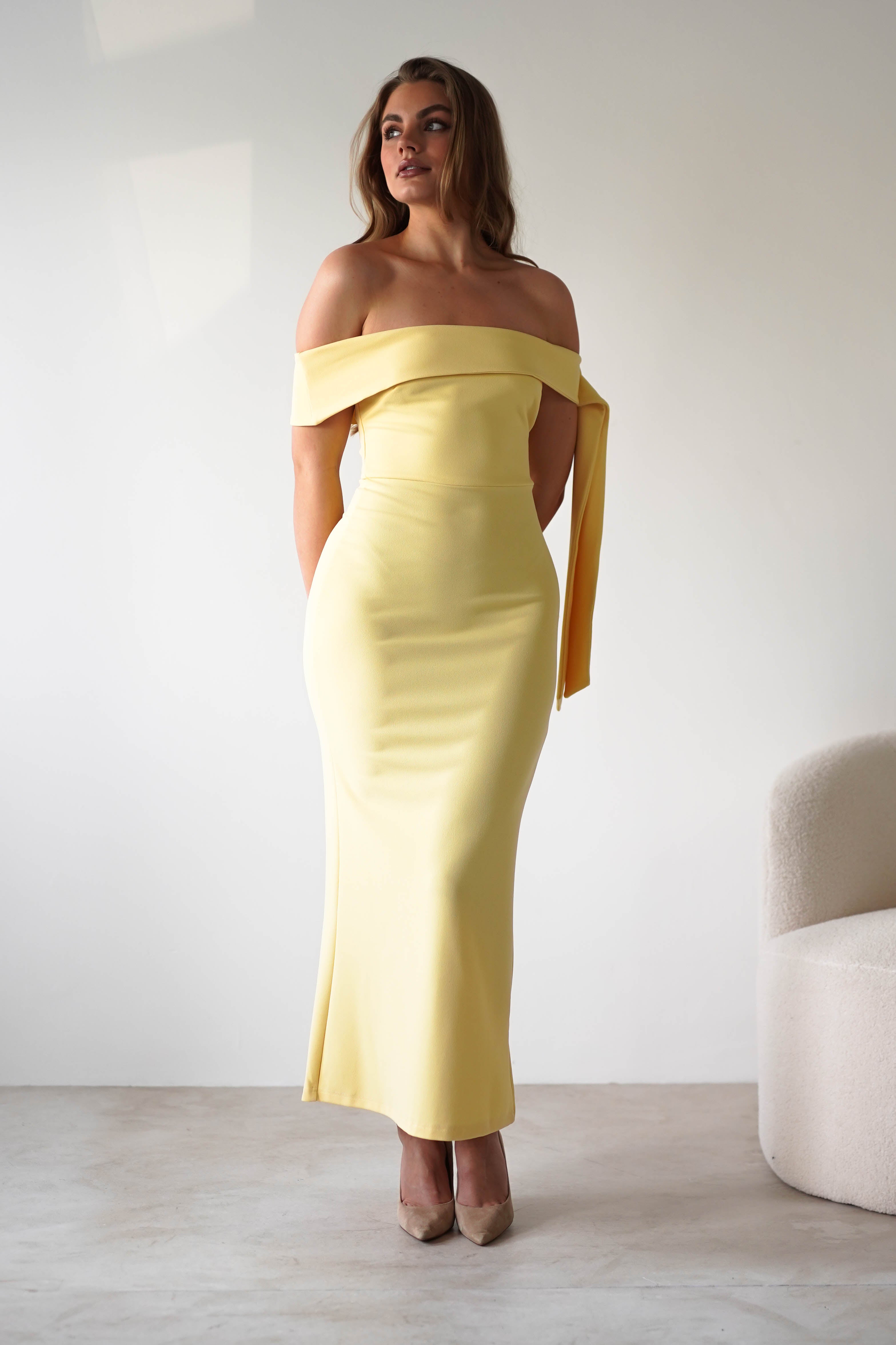 A woman stands in a minimalist room wearing an off-the-shoulder dress, perfect for a wedding guest. Her hair is in loose waves, high heels add elegance, and a cream-colored textured chair sits to the right.
