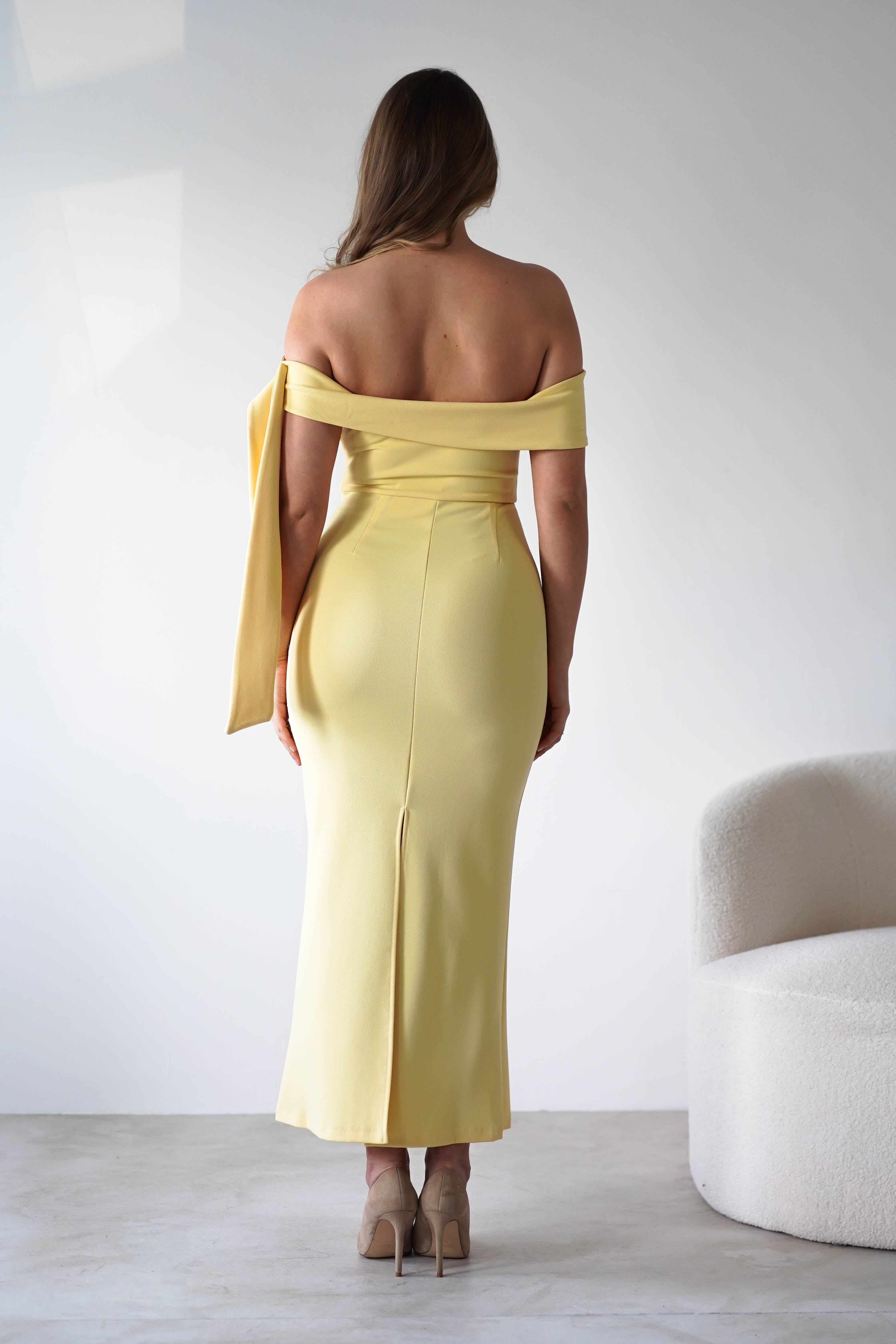 A woman stands facing away, wearing a fitted yellow off-the-shoulder dress and beige heels. With long brown hair, she looks ready as a chic wedding guest beside a white curved chair against a plain light-colored background.