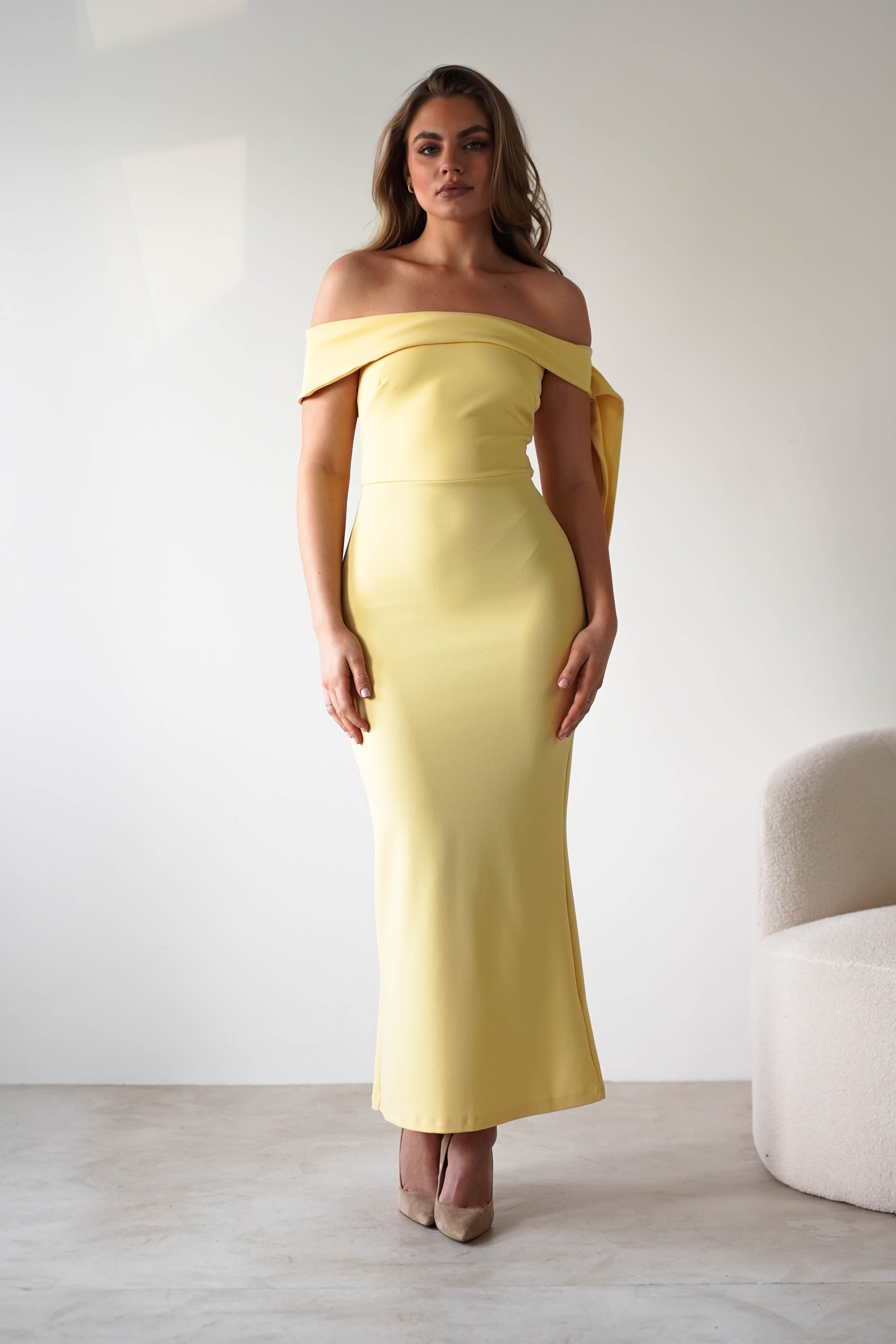 A woman stands indoors wearing a fitted, off-the-shoulder dress in yellow with beige heels. She poses in front of a plain white wall next to a curved, textured chair—an elegant look perfect for a wedding guest.