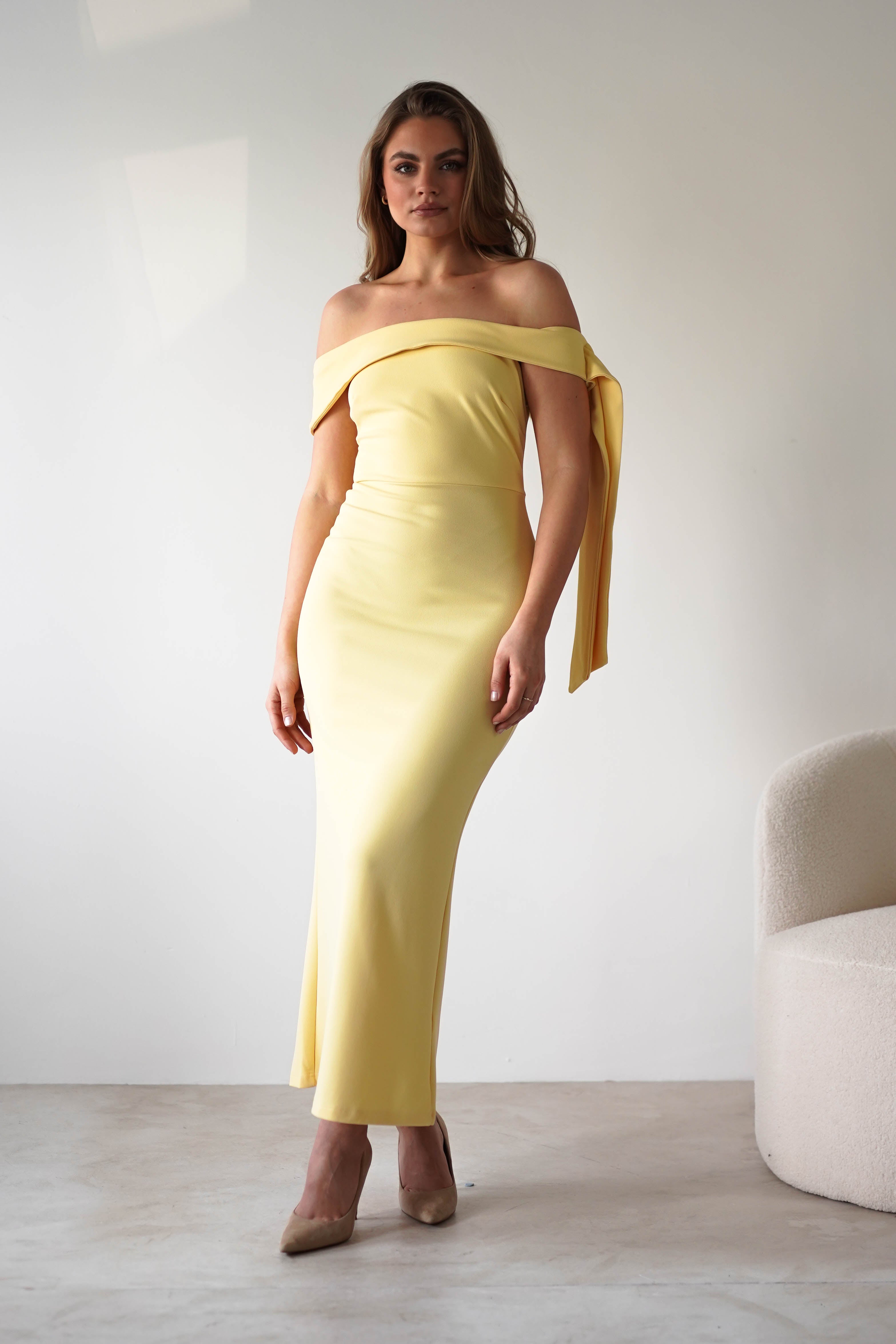Woman wearing a long, off-the-shoulder dress with a fitted silhouette and side tie detail—perfect for a wedding guest—standing indoors near a white textured chair against a plain light background.