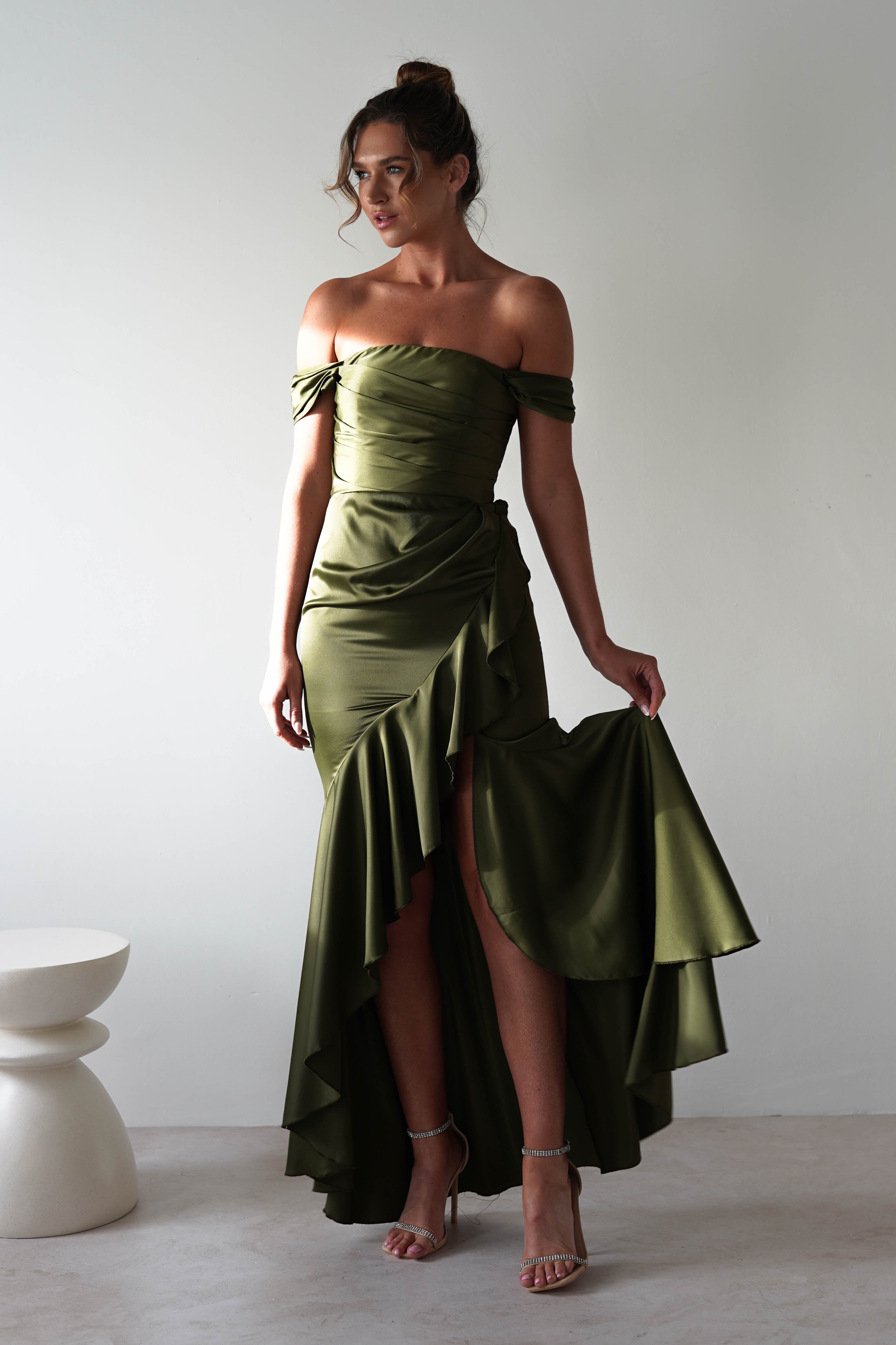 A woman in a chic, off-the-shoulder gown in olive green satin with a high-low hem and ruffles stands on a light floor against a white wall. She holds the side of her dress, highlighting strappy heels—a perfect ensemble for formal events with its modern flair and minimal decor.