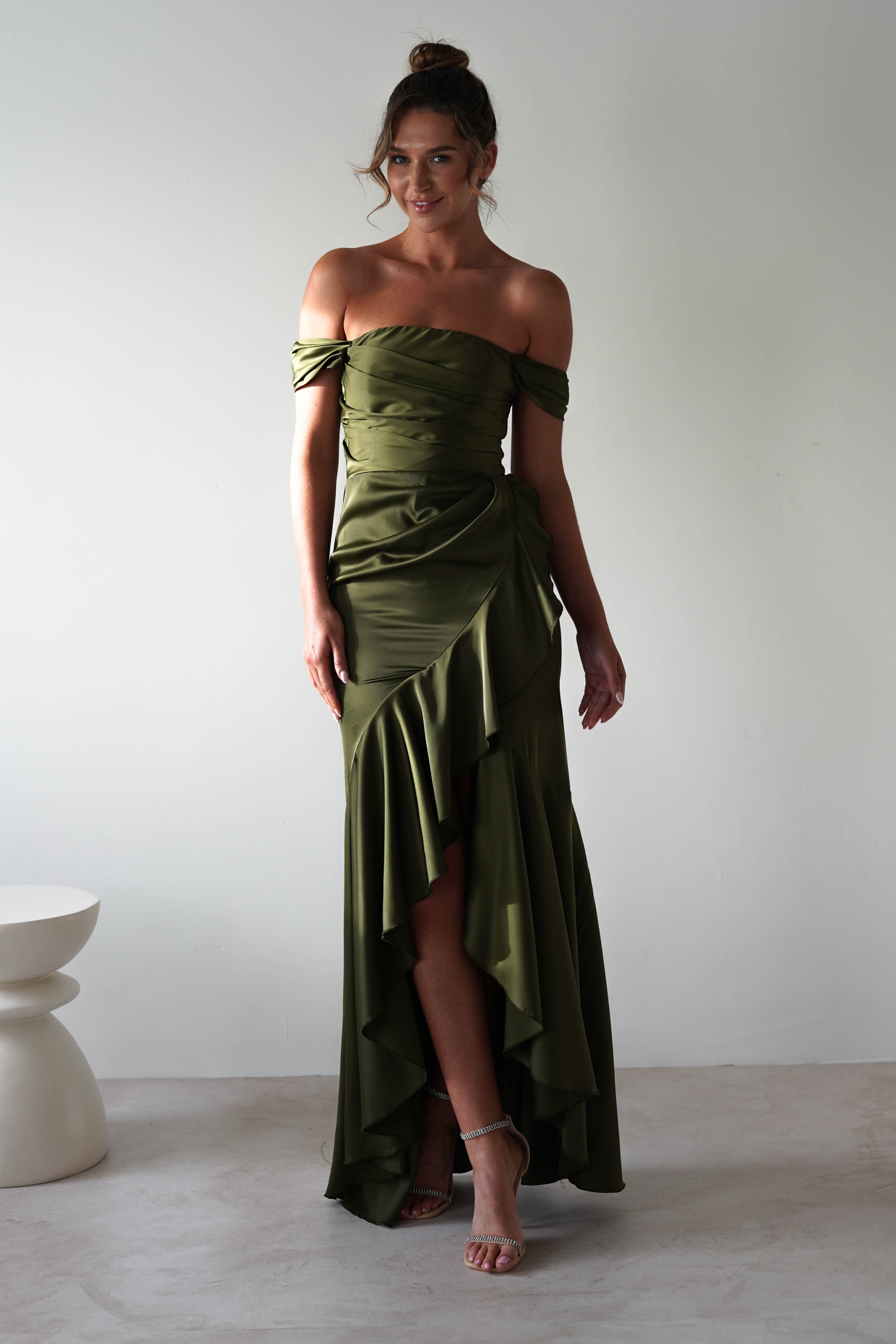 A person in an elegant off-the-shoulder gown made of soft satin stands against a plain white background. The olive green dress, ideal for formal events, features ruffled details and a high-low hemline. Theyre wearing heels with their hair styled in an updo, next to a small white stool.