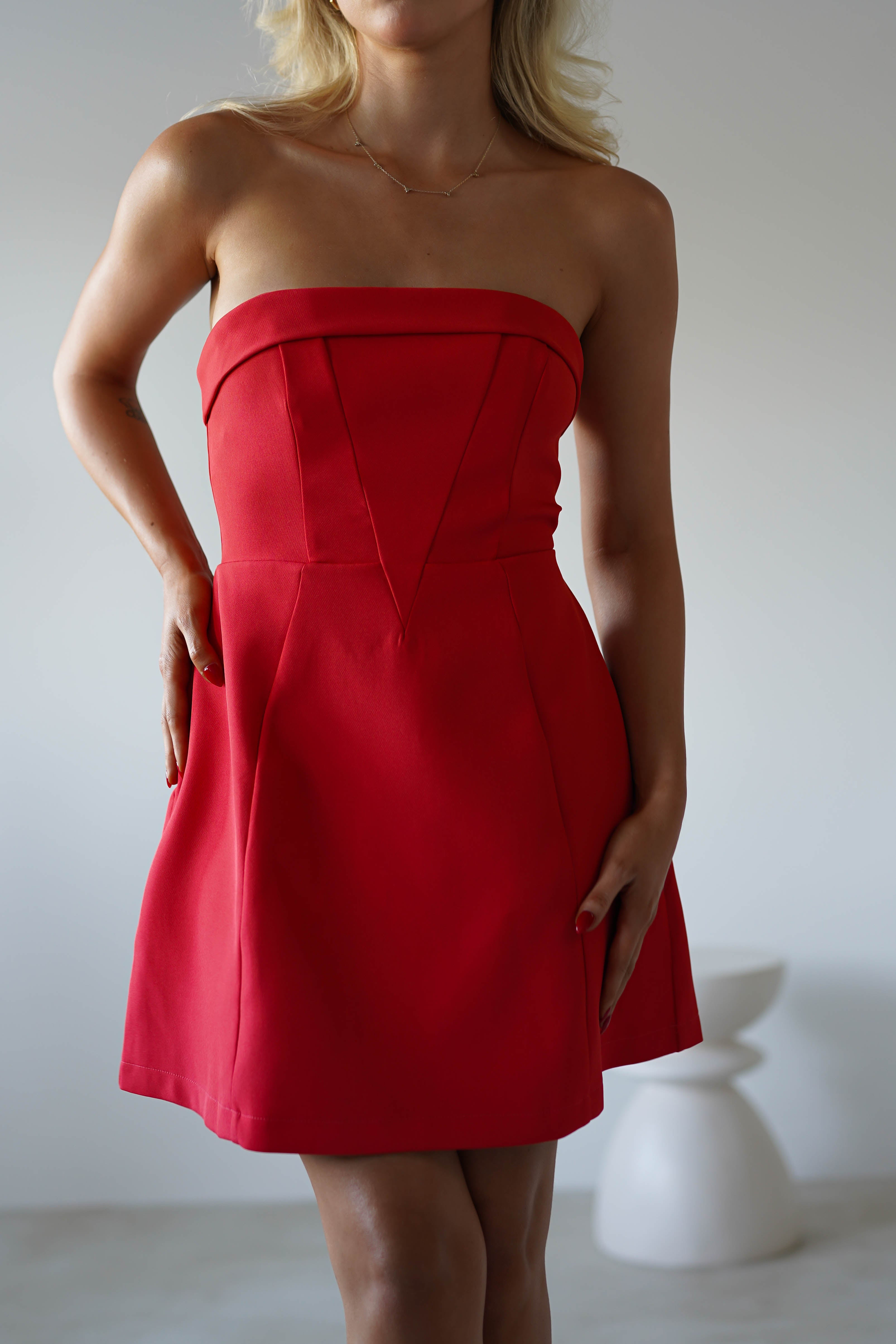 A woman wearing a strapless mini dress in fitted red stands indoors with one hand on her hip and the other by her side—a chic look perfect for graduates parties or as a stylish wedding guest. The background is neutral and minimalistic.