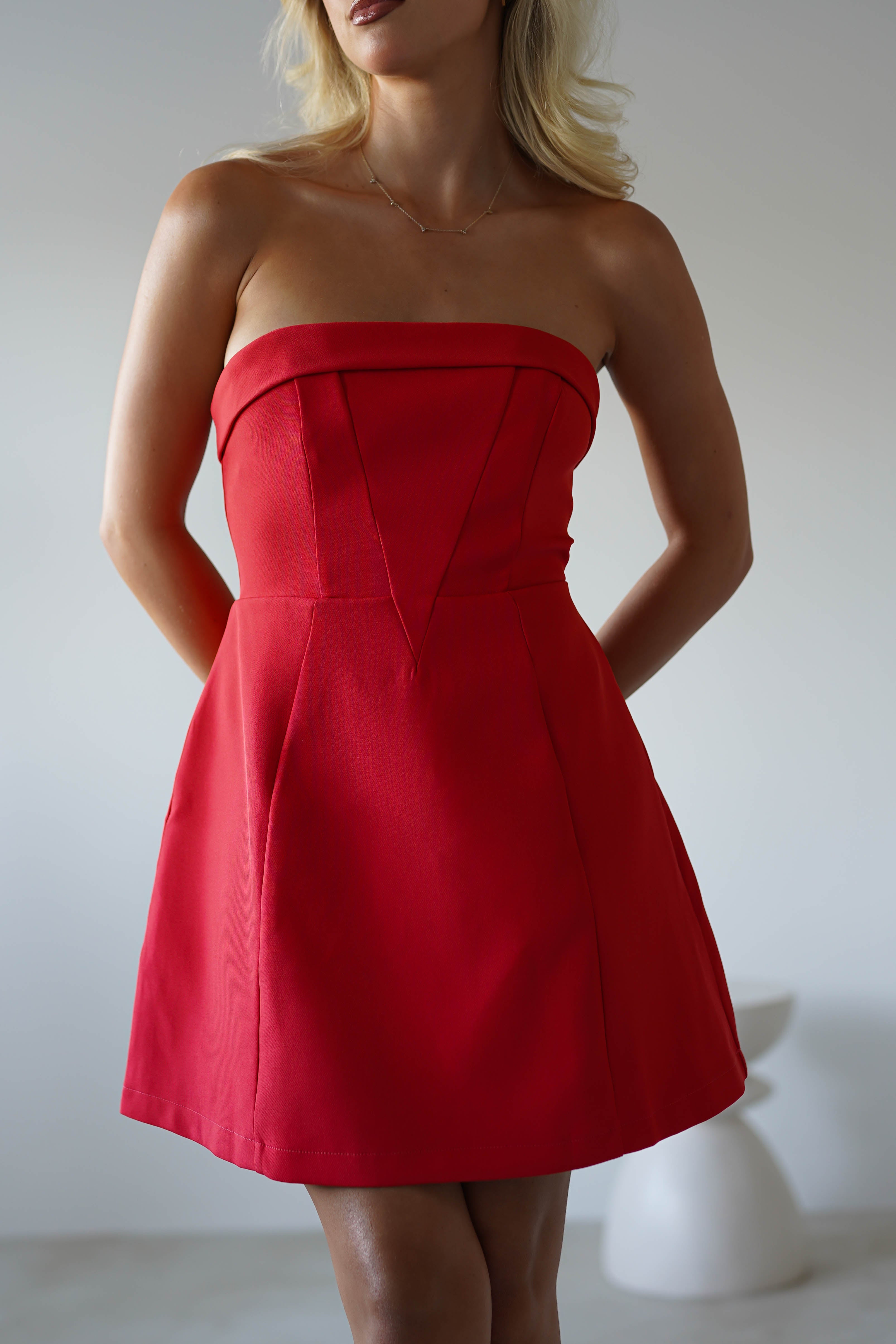 A woman wearing a strapless mini dress in vibrant red stands indoors with her hands behind her back. The softly blurred background and neutral lighting give this perfect look for graduates parties or as a stylish wedding guest.