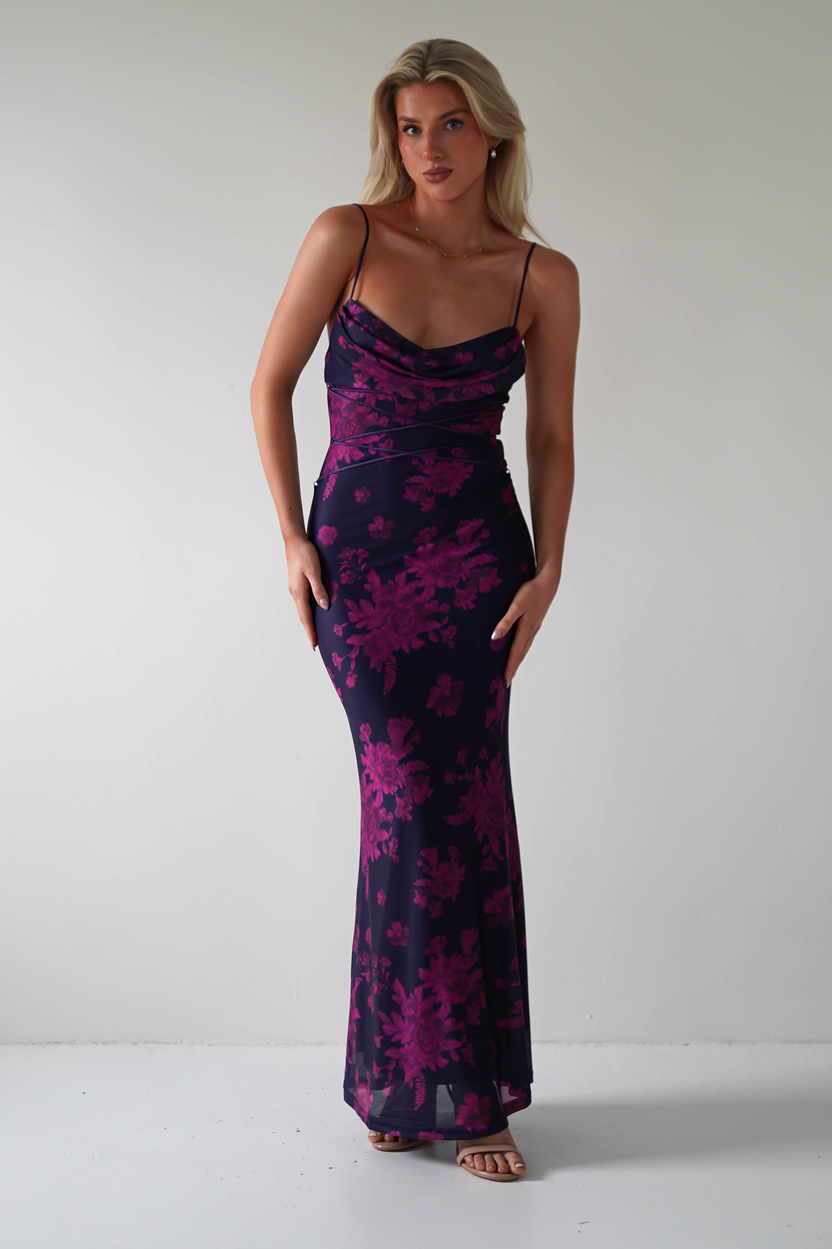 A woman poses confidently against a plain background, showcasing a stunning, form-fitting sleeveless navy dress adorned with pink floral patterns. The lace-up detail on the back adds an elegant touch for special occasions, while her strappy heels emphasize her poised stance.