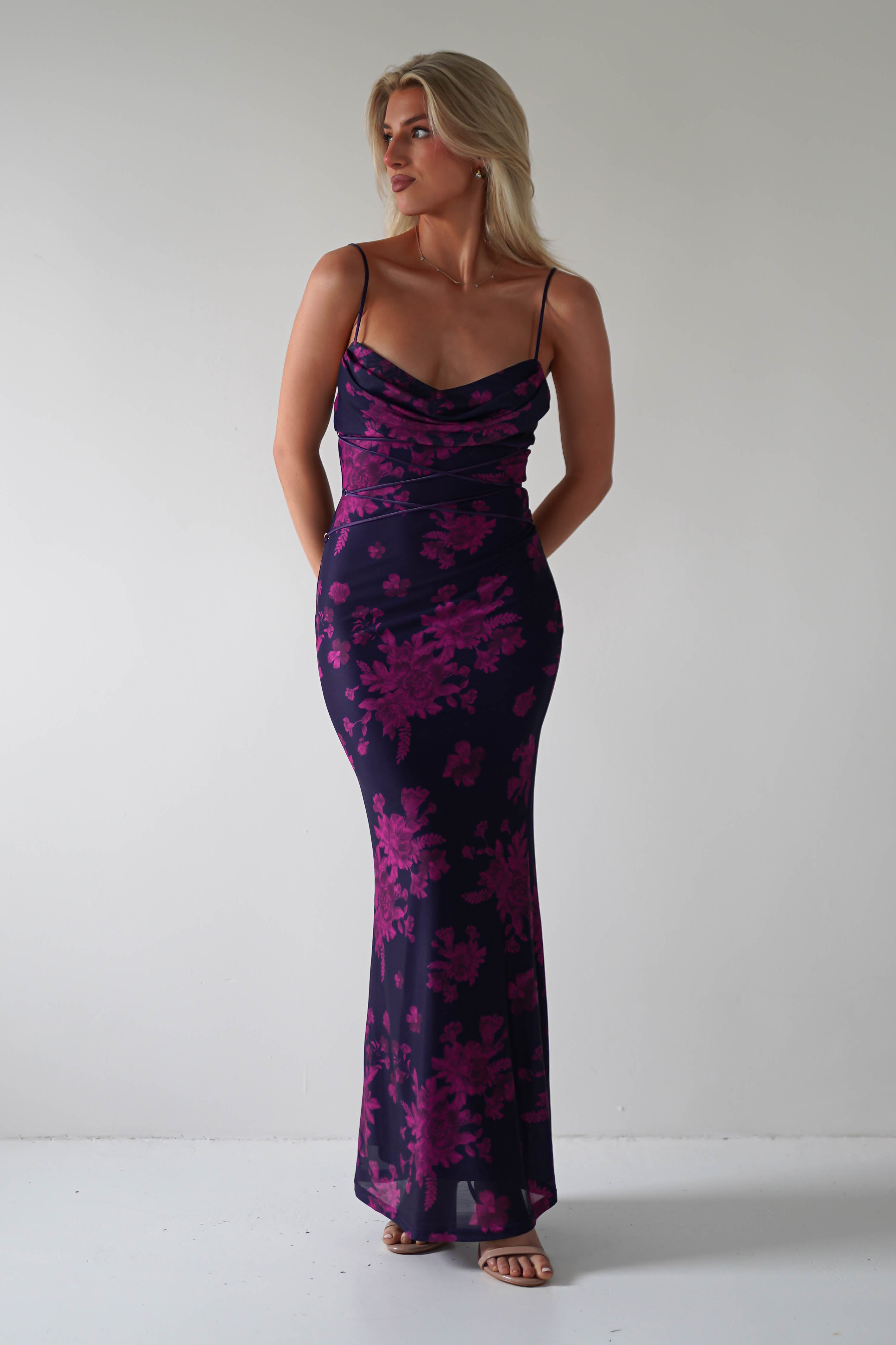A person is wearing a form-fitting, floor-length dress with thin straps and a floral pattern in shades of purple—a perfect choice for special occasions. The stretchy printed maxi elegantly hugs their frame as they pose against the plain, light-colored background.