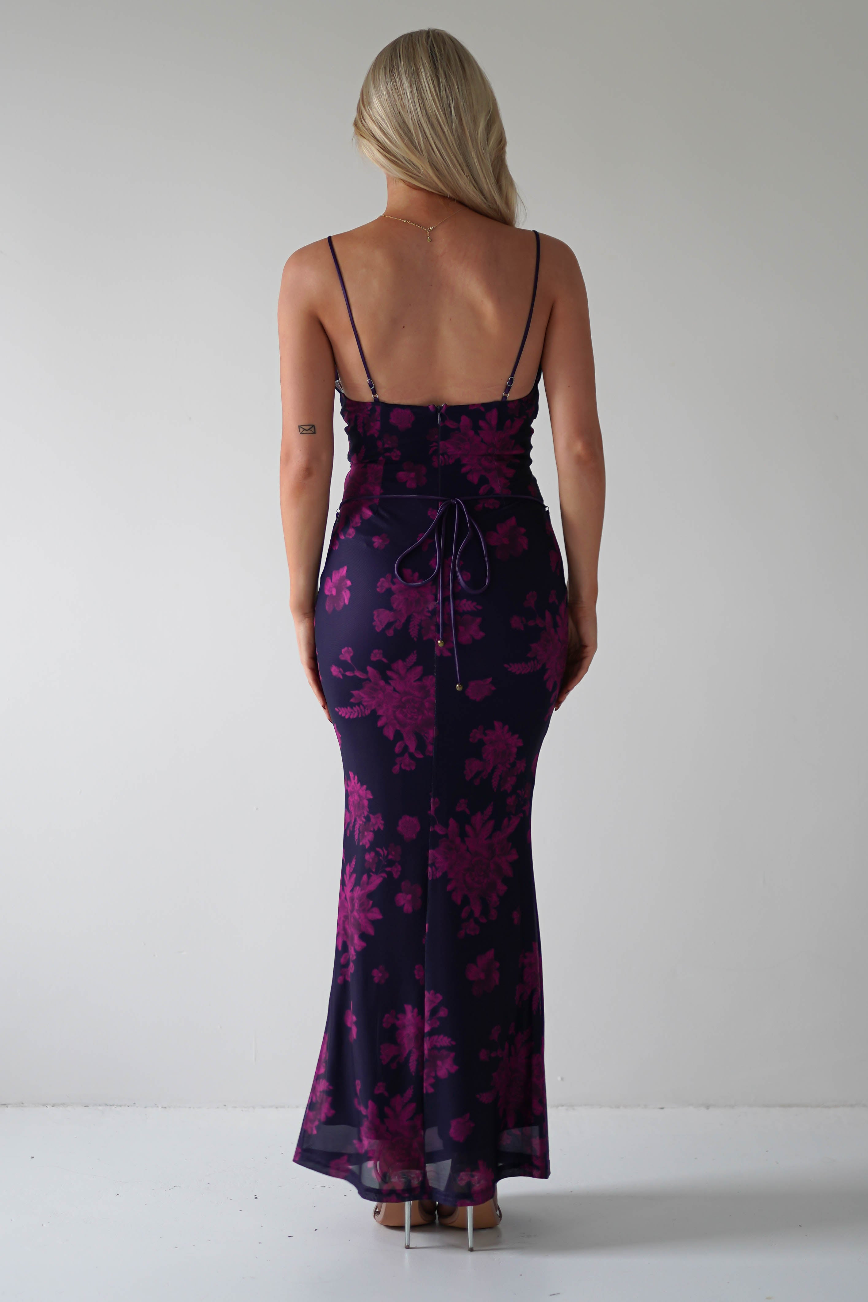 Rear view of a person with long blonde hair wearing a dark purple, floral-patterned dress featuring a lace-up detail and spaghetti straps, styled for special occasions. The dress ties elegantly at the back against a plain light gray background.