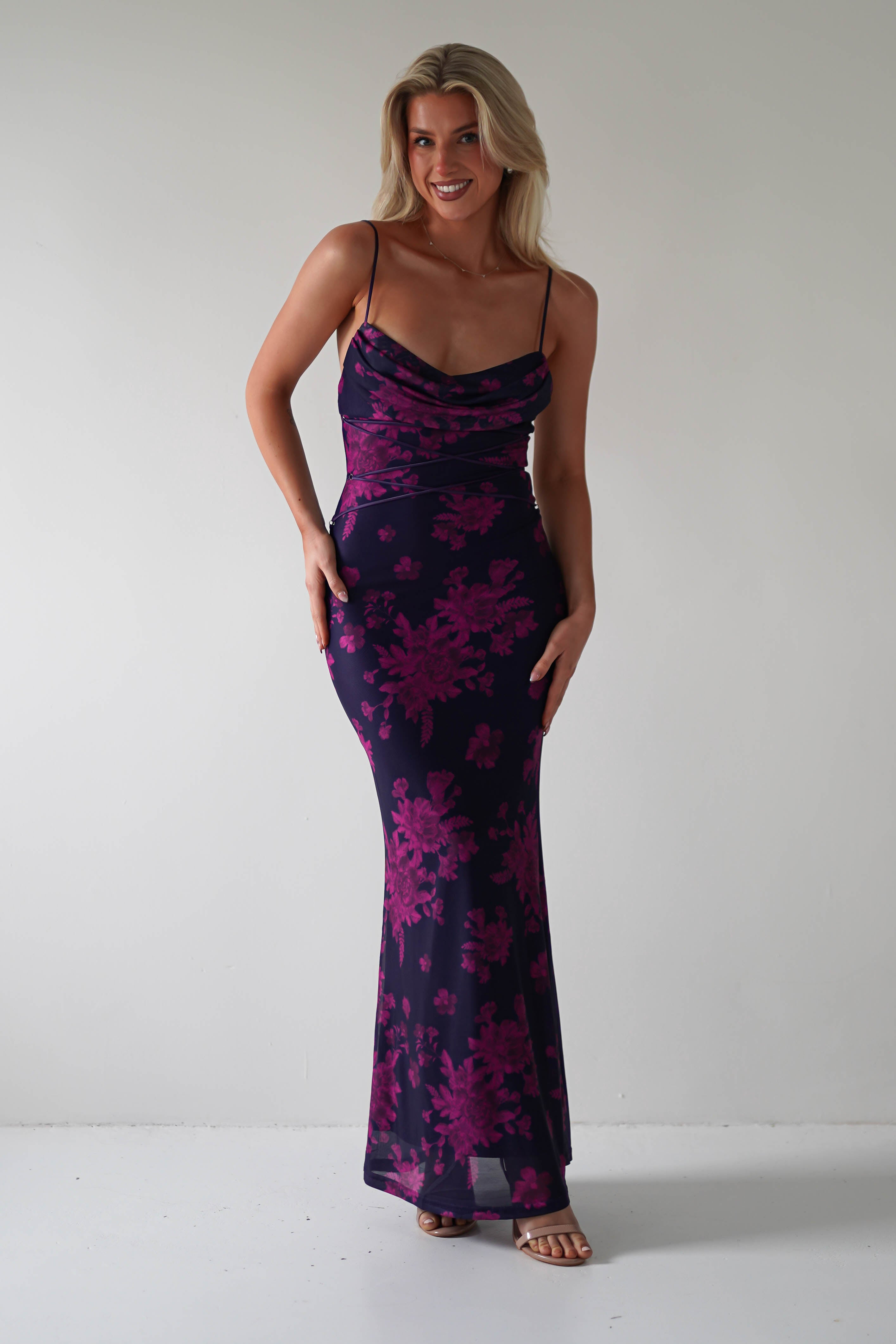 A person wearing a stretchy printed maxi dress with pink floral patterns stands smiling against a plain white background. Perfect for special occasions, the dark purple dress features thin straps and lace-up detail, complemented by stylish heels.