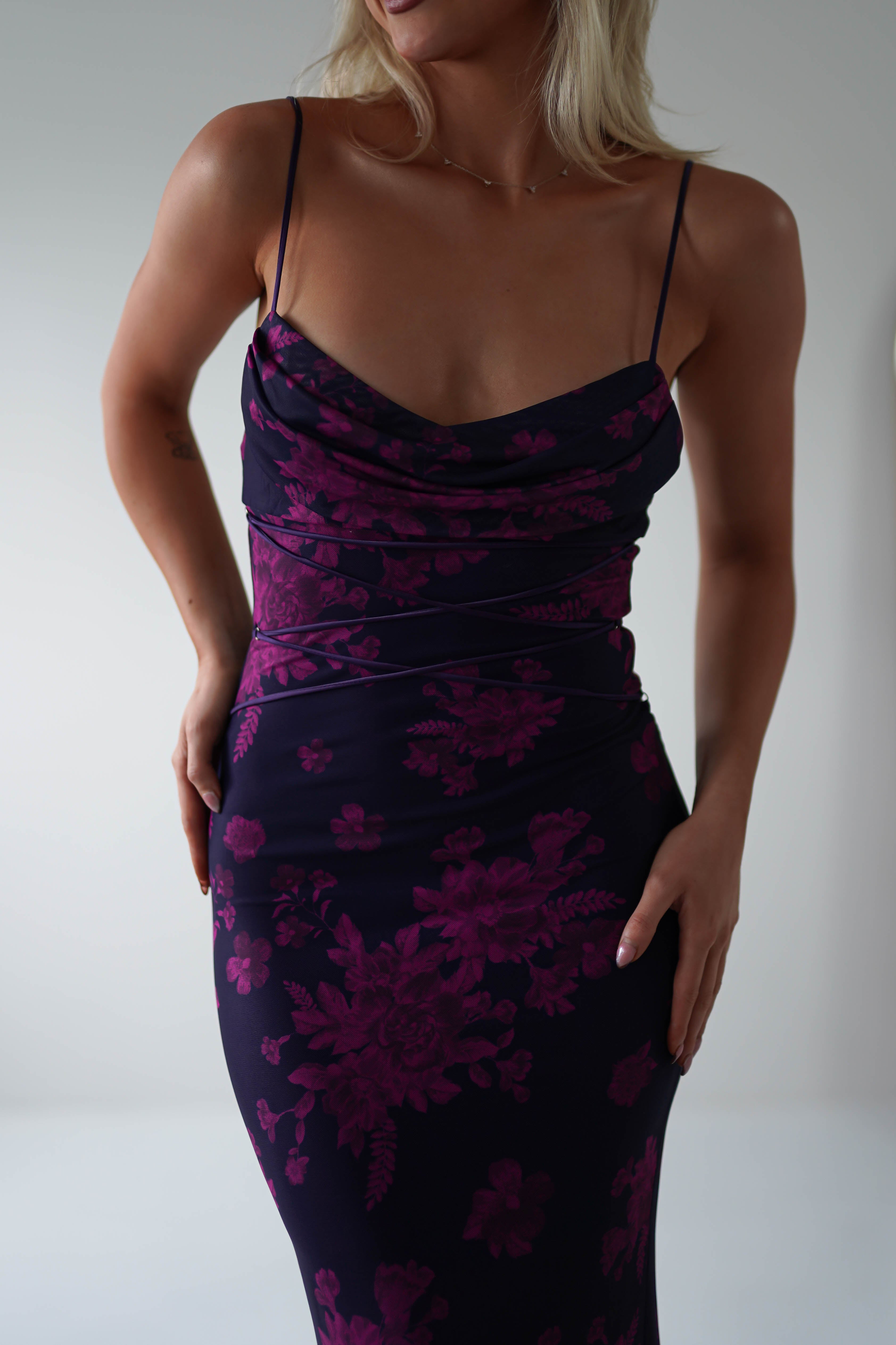 A person wearing a fitted, sleeveless dress with spaghetti straps, featuring a dark purple floral pattern on a black background. The stretchy printed maxi is perfect for special occasions. Theyre posing with one hand on their hip and the other touching the dress side. Face is not visible.