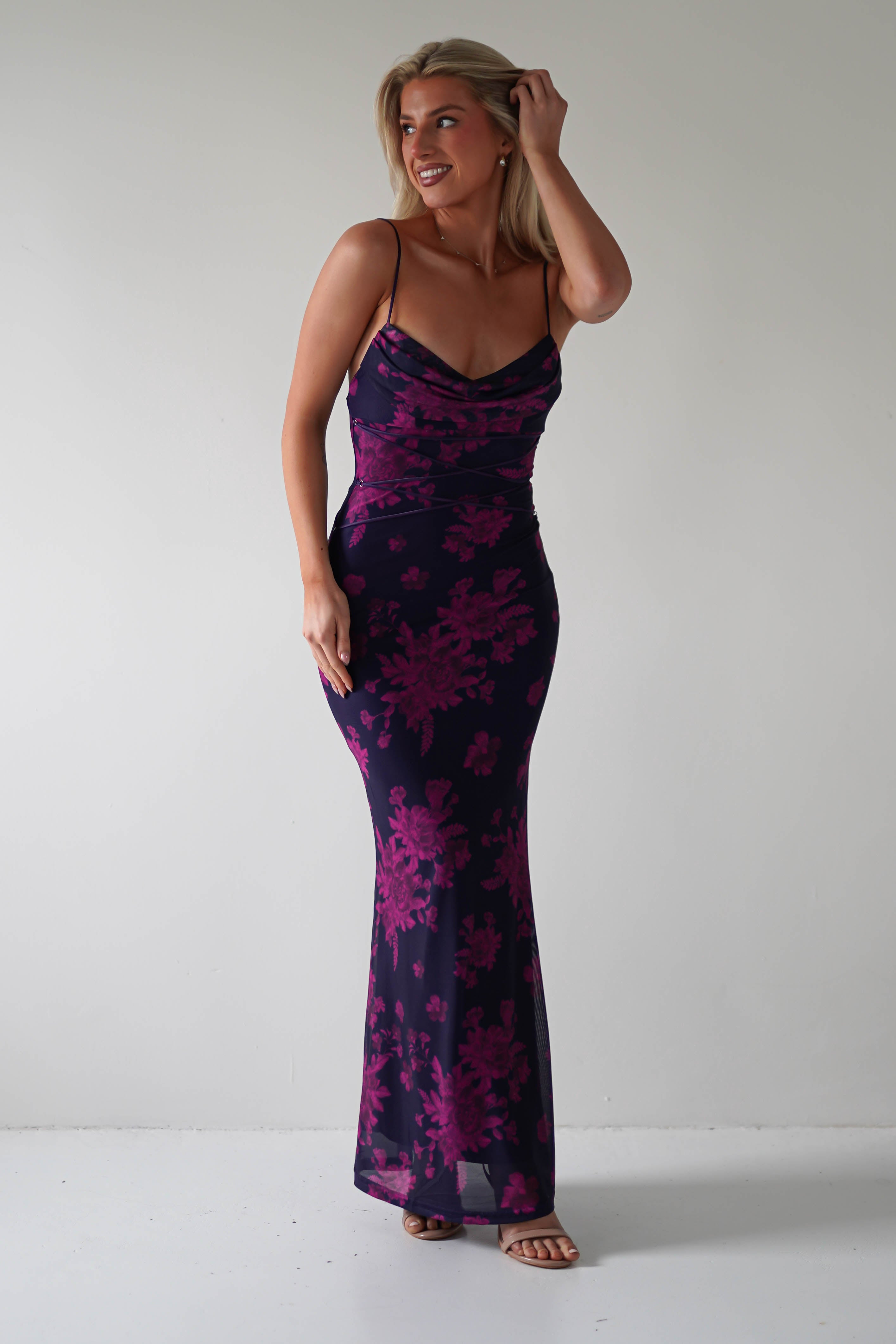 A person in a stretchy printed maxi with purple floral patterns stands smiling against a plain white background. They have their hand raised to their head, and the dress, perfect for special occasions, is complemented by strappy black heels.