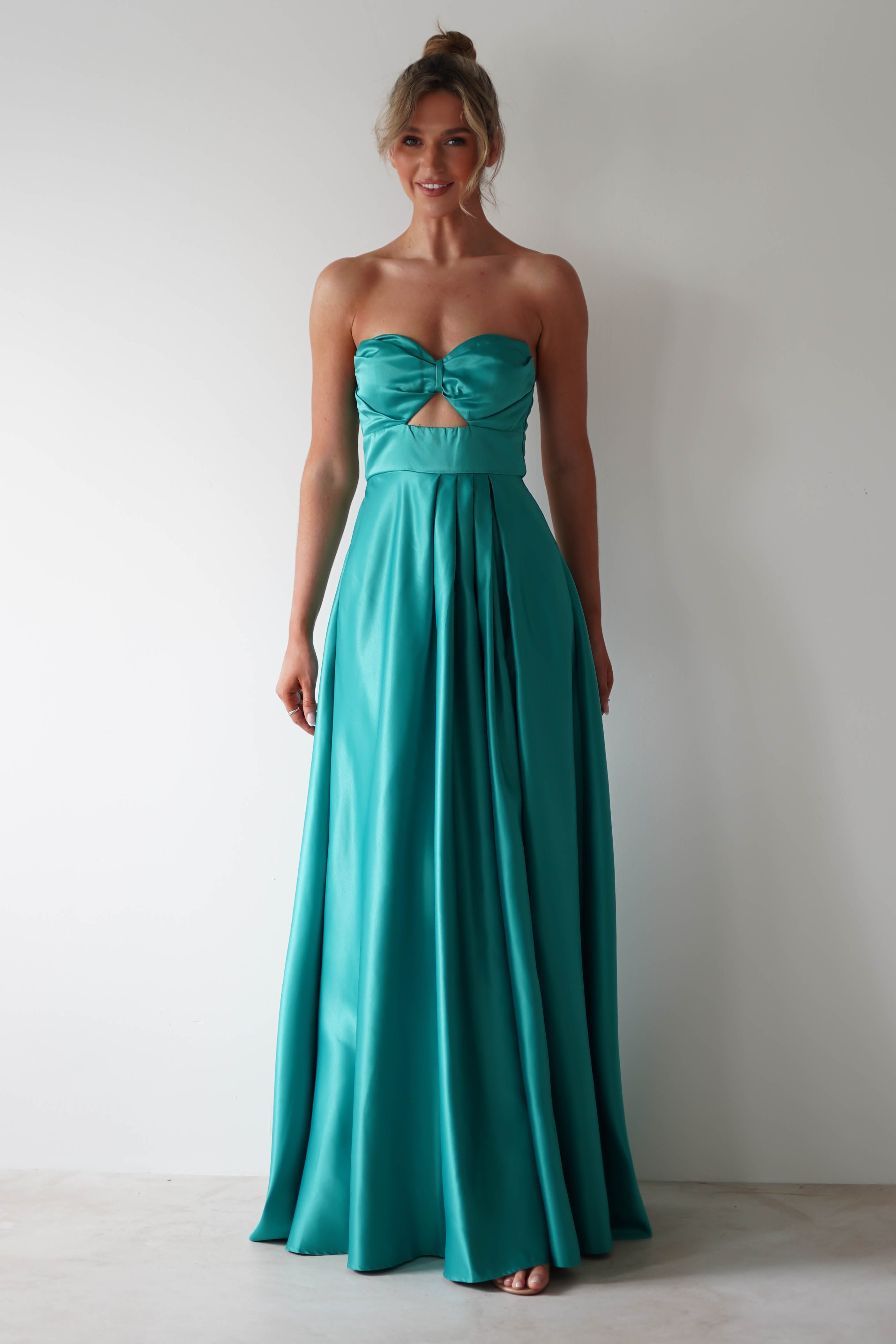 A woman wearing a strapless, teal, floor-length satin prom dress with a cut-out detail at the bust and a thigh high slit stands against a plain white background, smiling slightly.