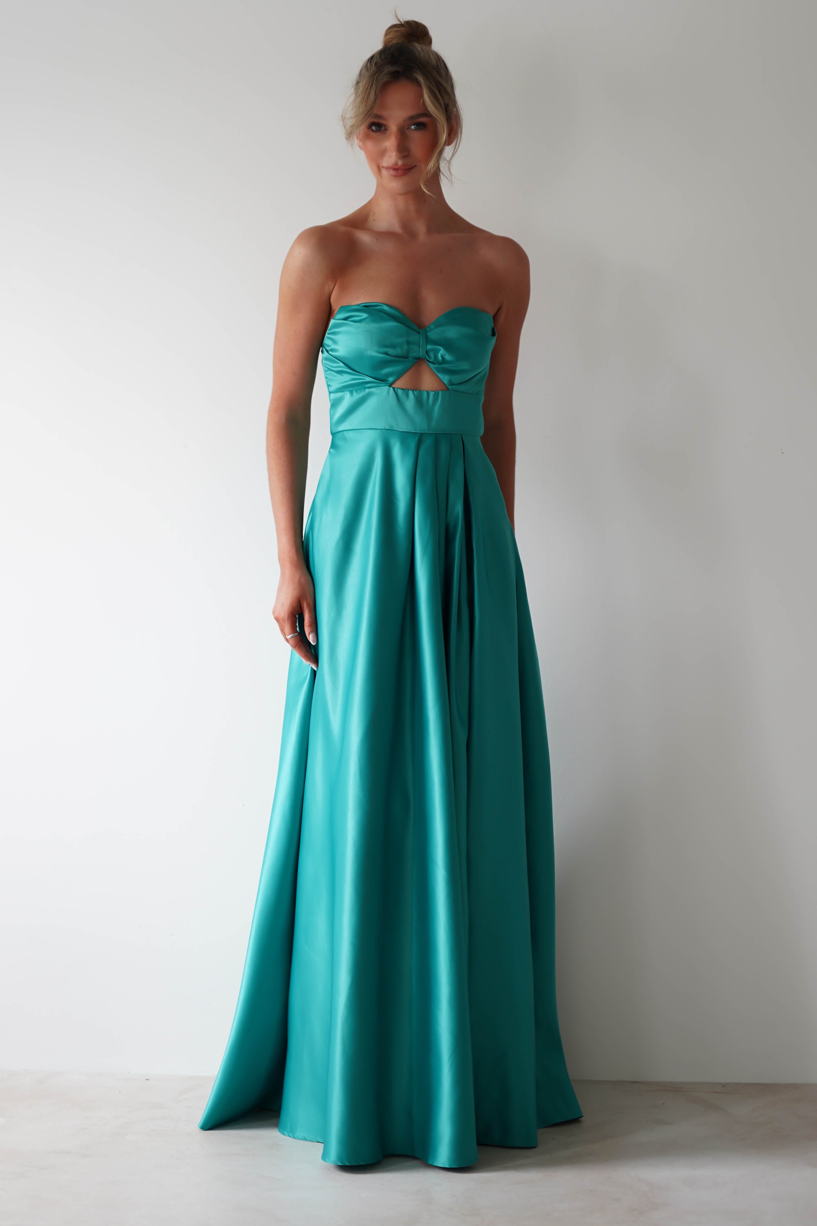 A woman stands against a plain light background wearing a strapless, floor-length prom dress in teal satin, featuring a front cutout, sash detail at the waist, and a daring thigh high slit.