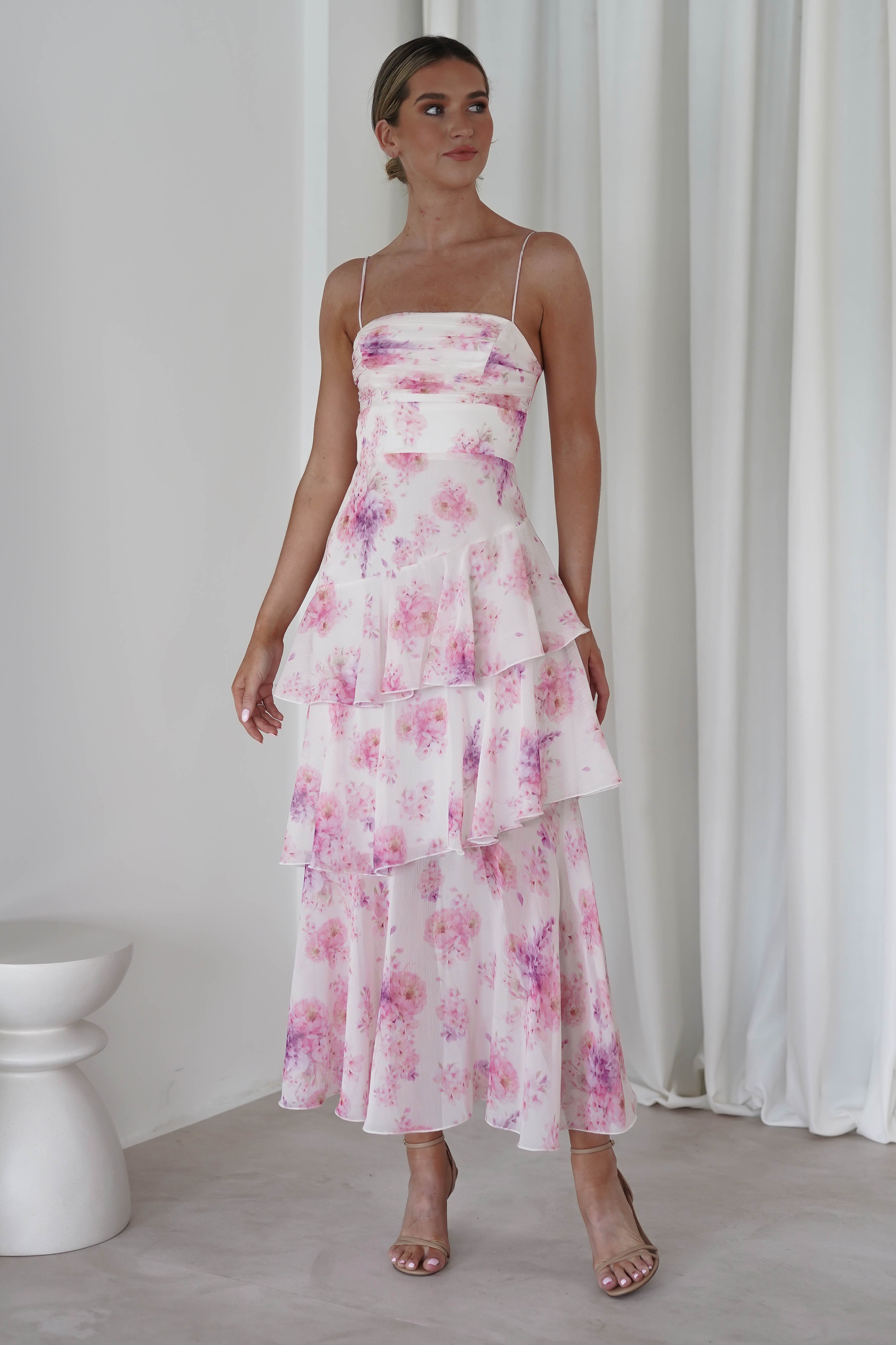 A woman in a pink floral midi with purple patterns stands indoors. The wedding guest dress features a layered ruffle design. She is wearing strappy heels, with her hair tied back and a neutral-colored curtain in the background.