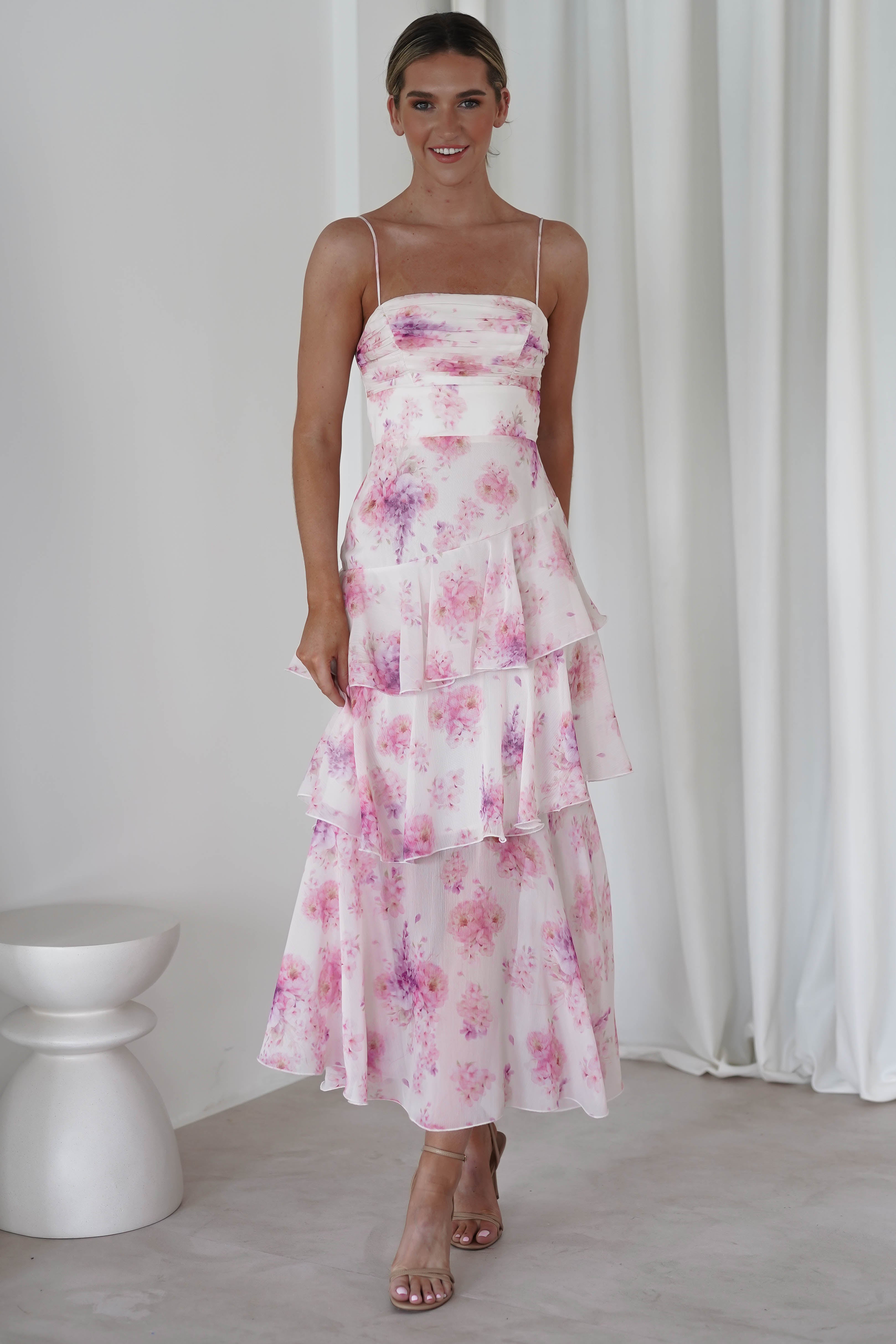 A woman in a pink floral midi dress, perfect for a wedding guest, stands elegantly in a light room. Her heels complete the look, with her hair neatly pulled back. The background showcases white curtains and a minimalist stool.