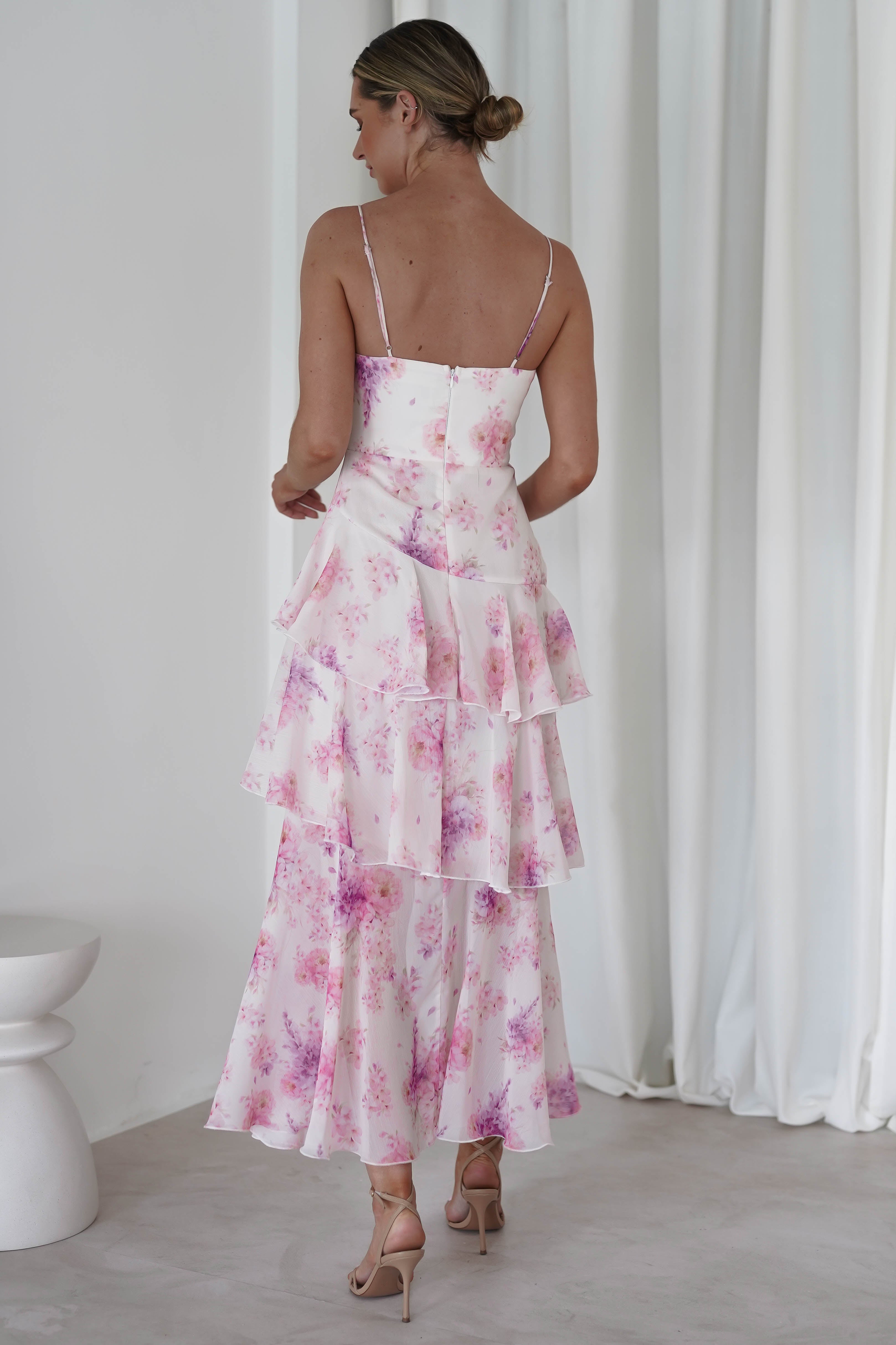A woman in a pink floral midi dress with tiered layers and thin straps stands with her back to the camera. Her chiffon wedding guest dress pairs elegantly with high heels, while her hair is styled in a chic bun. White curtains and a stool provide a simple backdrop.