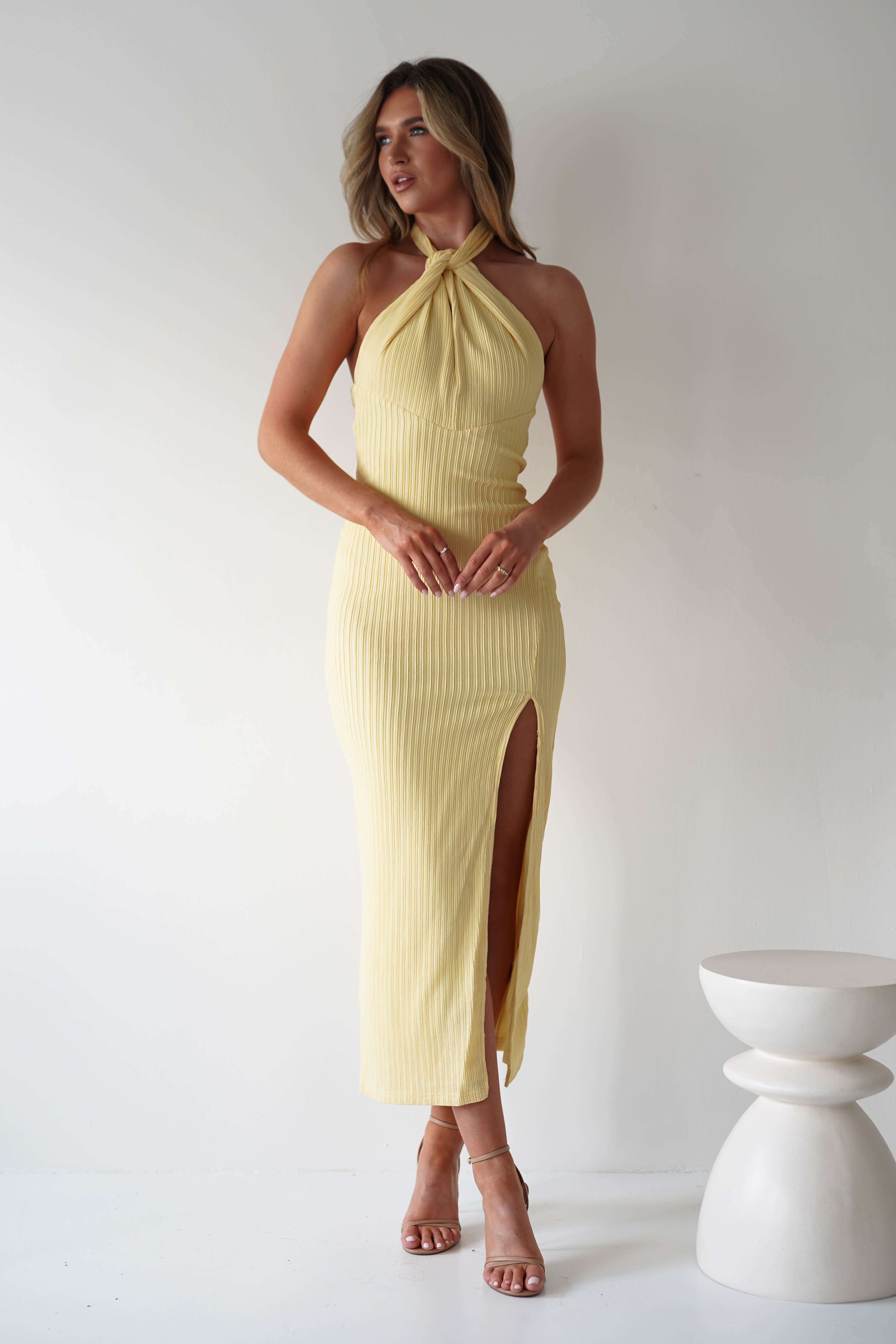 Woman poses in a sleeveless, light yellow textured midaxi dress with halterneck ties and a side slit. She wears strappy heels and stands next to a modern white pedestal, set against a plain white background—perfect for special occasions.