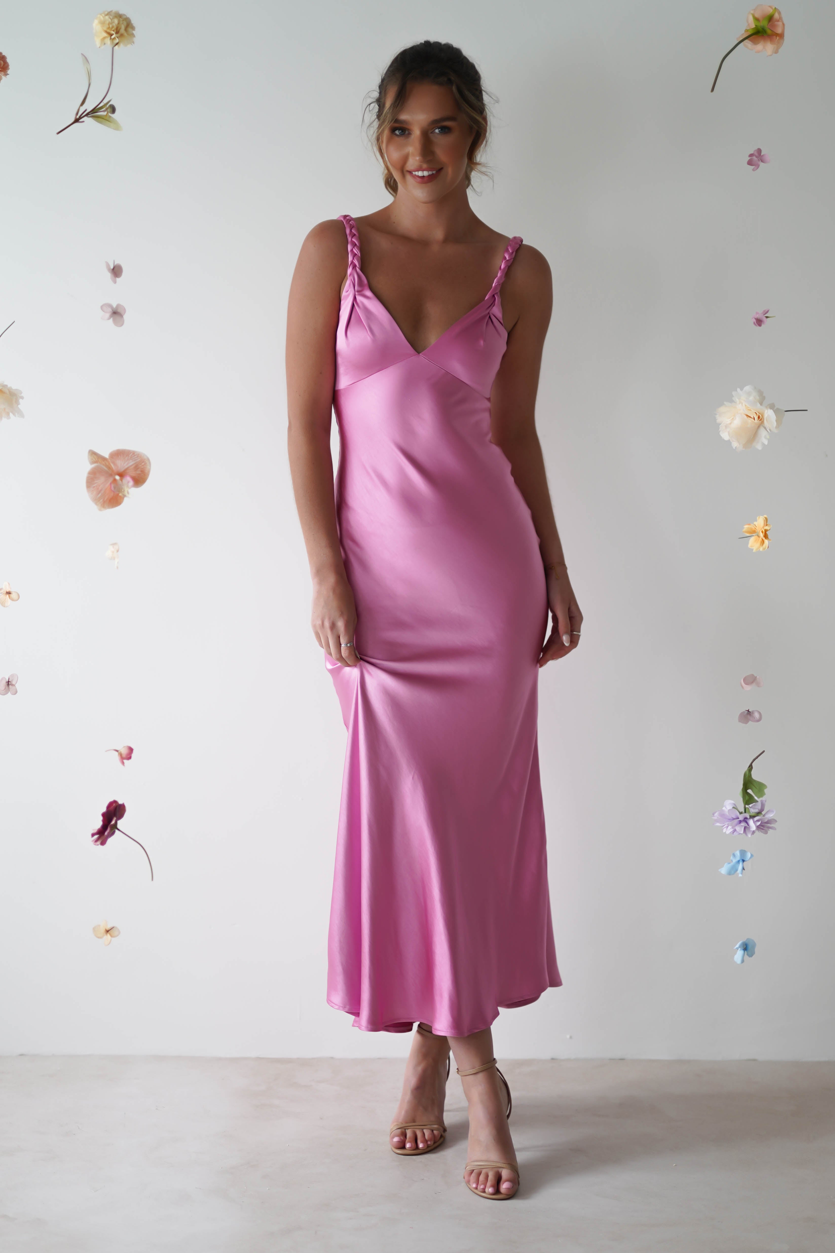 A woman in a pink satin maxi dress with adjustable straps stands against a white backdrop adorned with floating flowers. As a perfect wedding guest attire, she lightly holds the hem of her dress, accentuating her strappy heels.