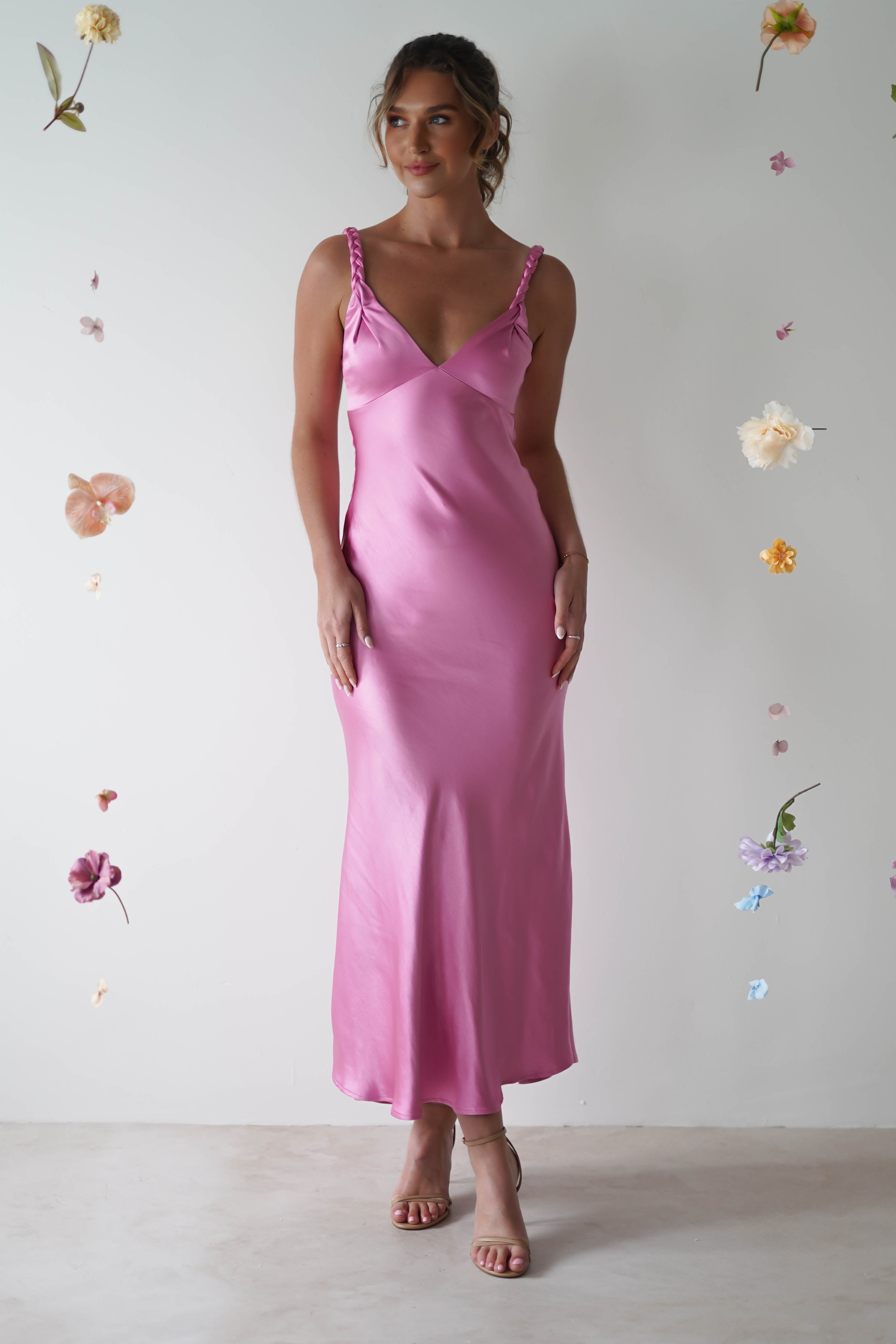A woman in a pink satin maxi dress with adjustable straps stands against a plain backdrop adorned with floating flowers. Her hair is styled up, and she gazes slightly to the side, hands relaxed by her sides—an elegant choice for any wedding guest.