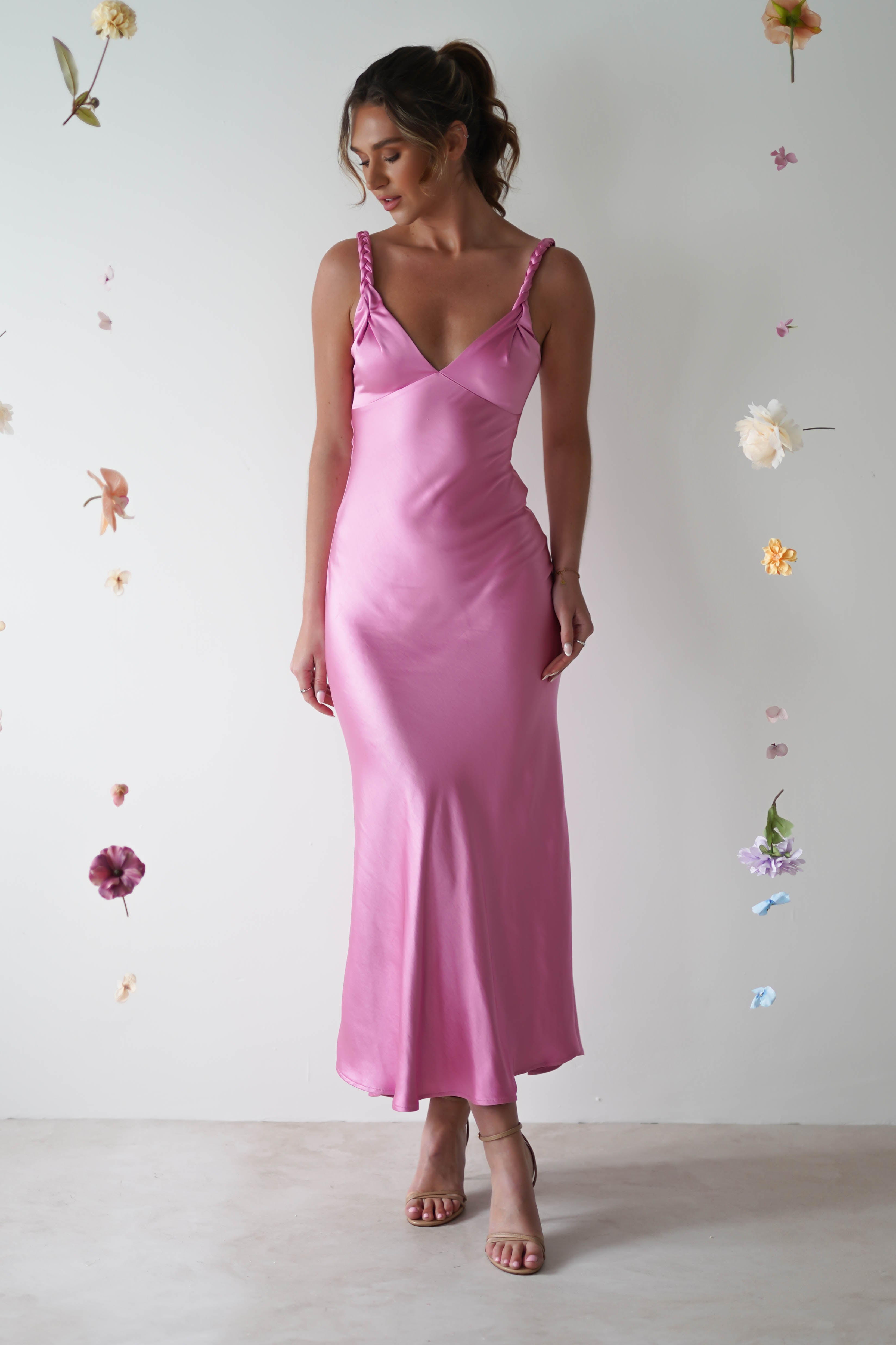 A woman in a pink satin maxi dress with adjustable straps stands against a white background adorned with floating, assorted flowers. Her hair is styled in an updo, complemented by simple jewelry, crafting an elegant and delicate scene perfect for a wedding guest.
