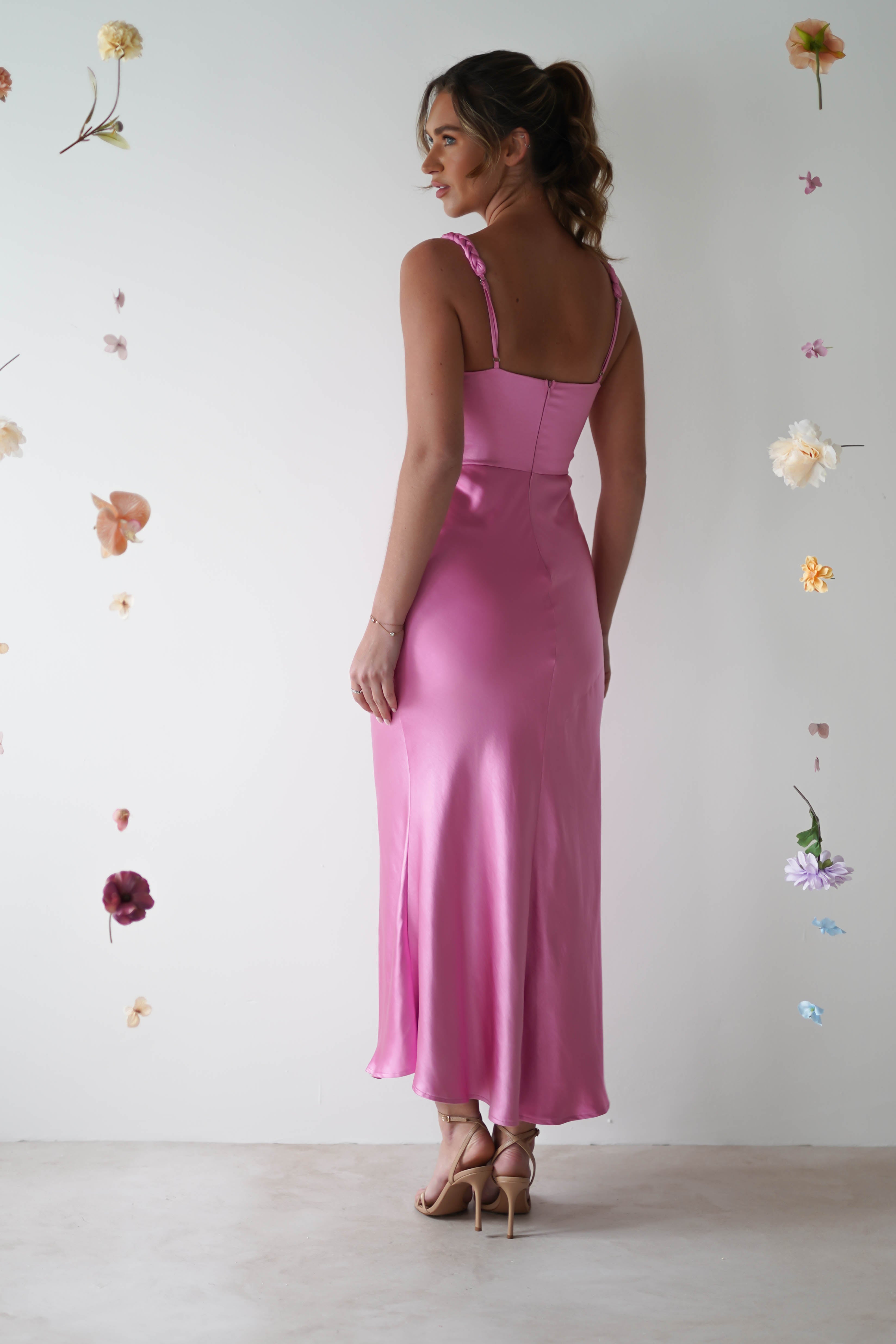 A woman stands gracefully in a silky pink satin maxi dress, facing away. The dress is long and form-fitting with adjustable straps. She is surrounded by floating flowers against a plain background, creating an ethereal wedding guest effect.