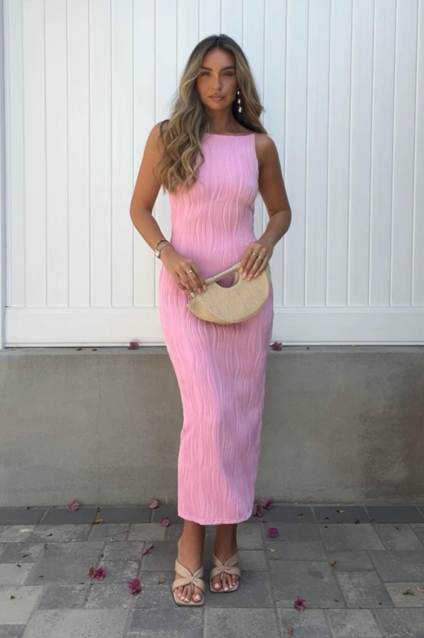 A woman poses in front of a white wall, gracing a sleeveless pink textured midaxi dress and sandals. She holds a semi-circular straw clutch, her long wavy hair cascading down. Pink petals are scattered on the ground, adding a touch of charm for special occasions.