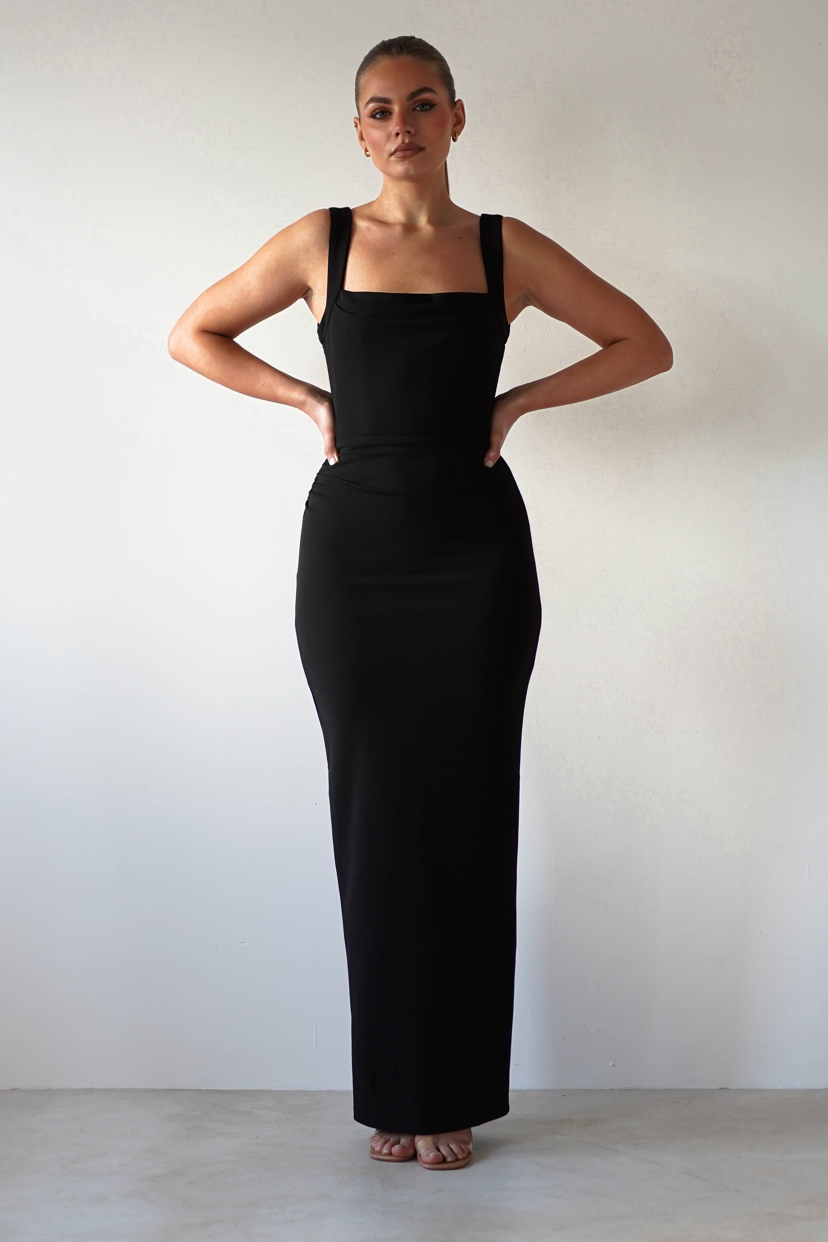A person stands confidently against a plain background, wearing a glamorous bodycon fit black dress with shoulder straps. Their hair is styled up, and they have their hands on their hips, conveying a poised and elegant stance.