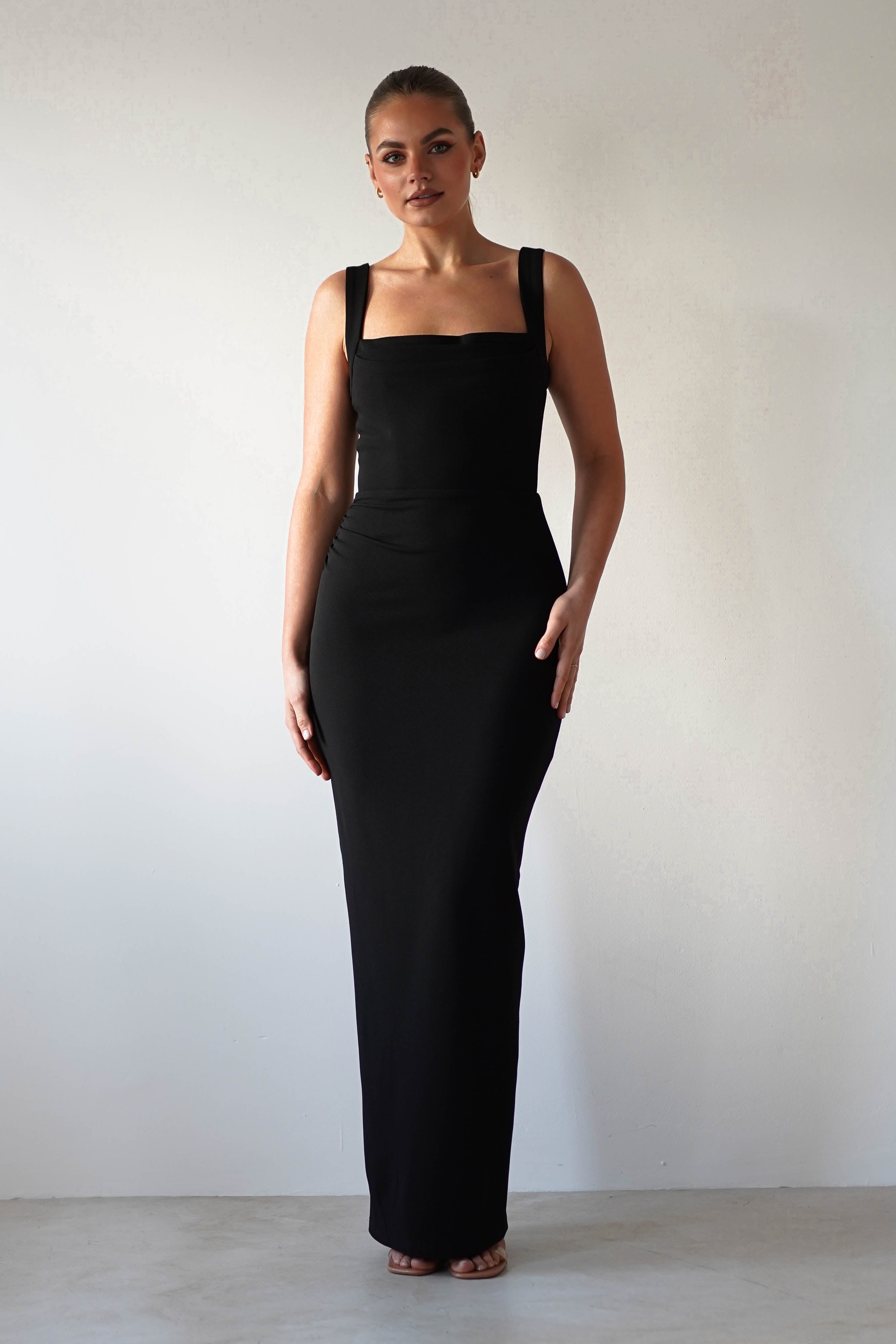 A person stands against a plain background wearing a glamorous, form-fitting, sleeveless black dress with thick straps. The square neck design complements their neutral expression, with one hand on their hip. The lighting highlights the dresss sleek silhouette and bodycon fit beautifully.