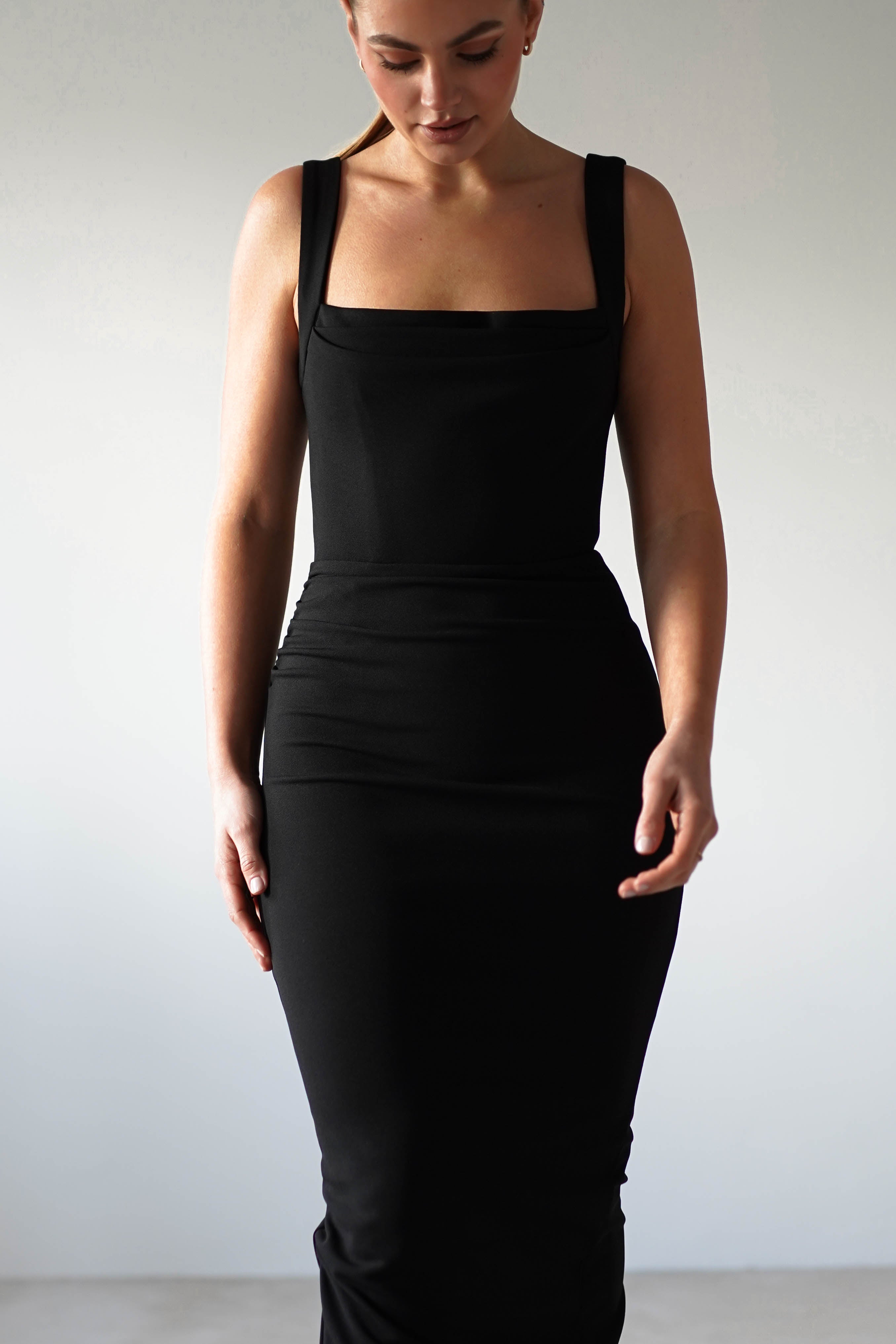 Person wearing a glamorous, fitted black square neck dress stands against a plain background. The dress, with its bodycon fit and wide straps, elegantly falls below the knees. The individuals hands rest relaxed by their sides, exuding effortless sophistication.