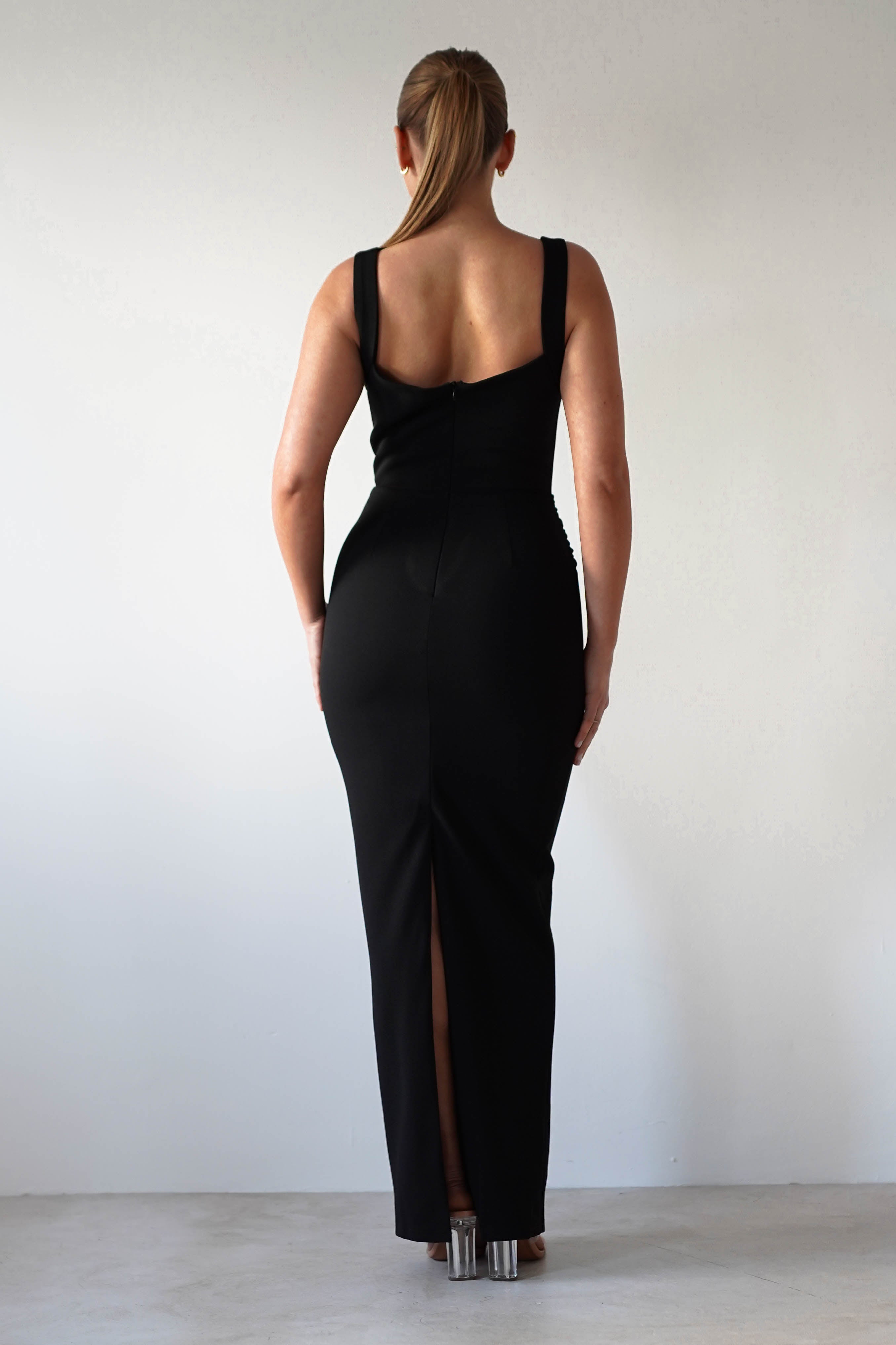 A woman stands facing away, wearing a glamorous black evening gown with a slit at the back. The dress showcases a bodycon fit that highlights her silhouette. Her hair is in a ponytail, and she wears clear-heeled shoes against a plain white wall backdrop.
