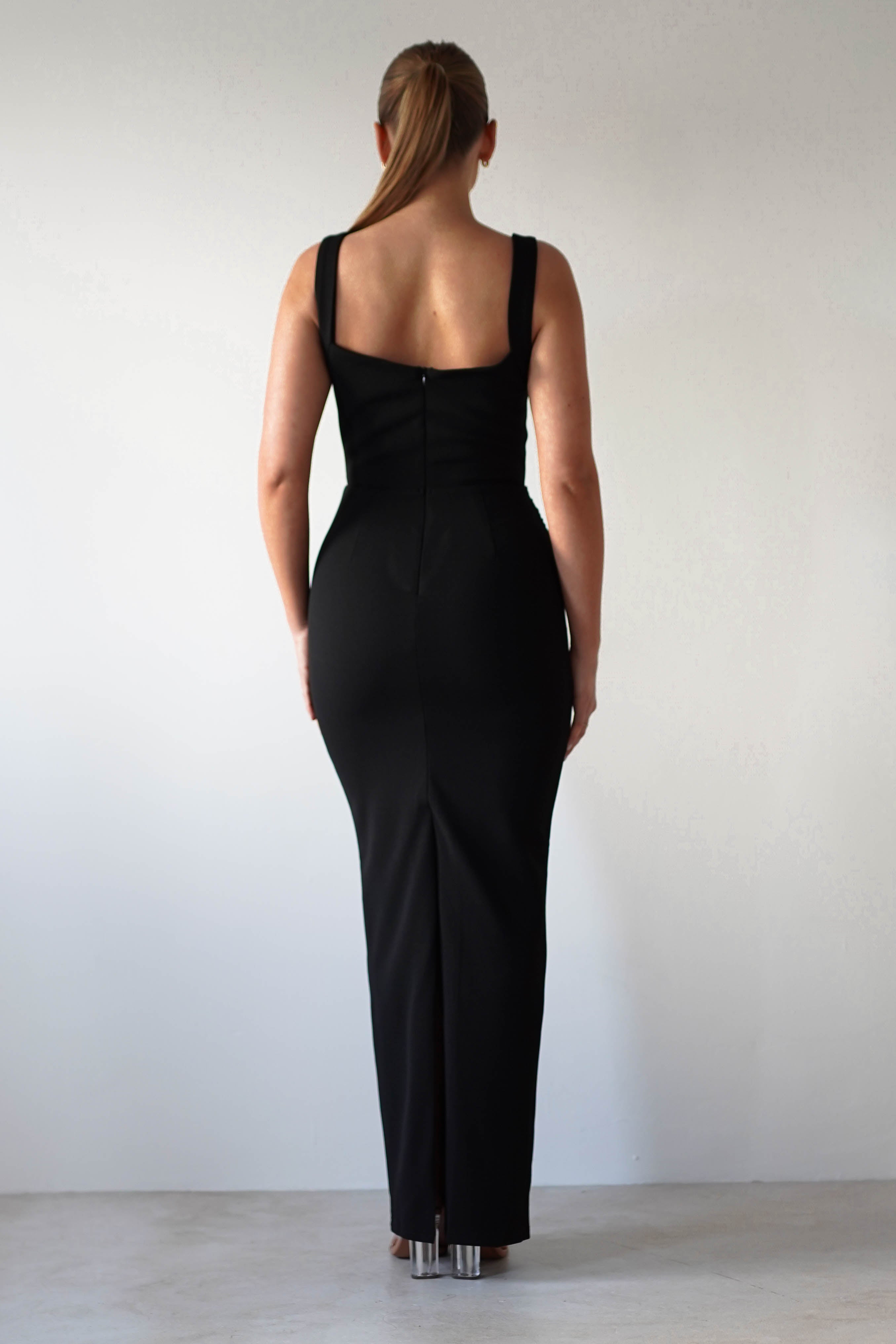 Woman in a long, black, square neck dress standing with her back to the camera against a plain white background. She has her hair pulled back in a ponytail, exuding a glamorous charm with the bodycon fit accentuating her silhouette.