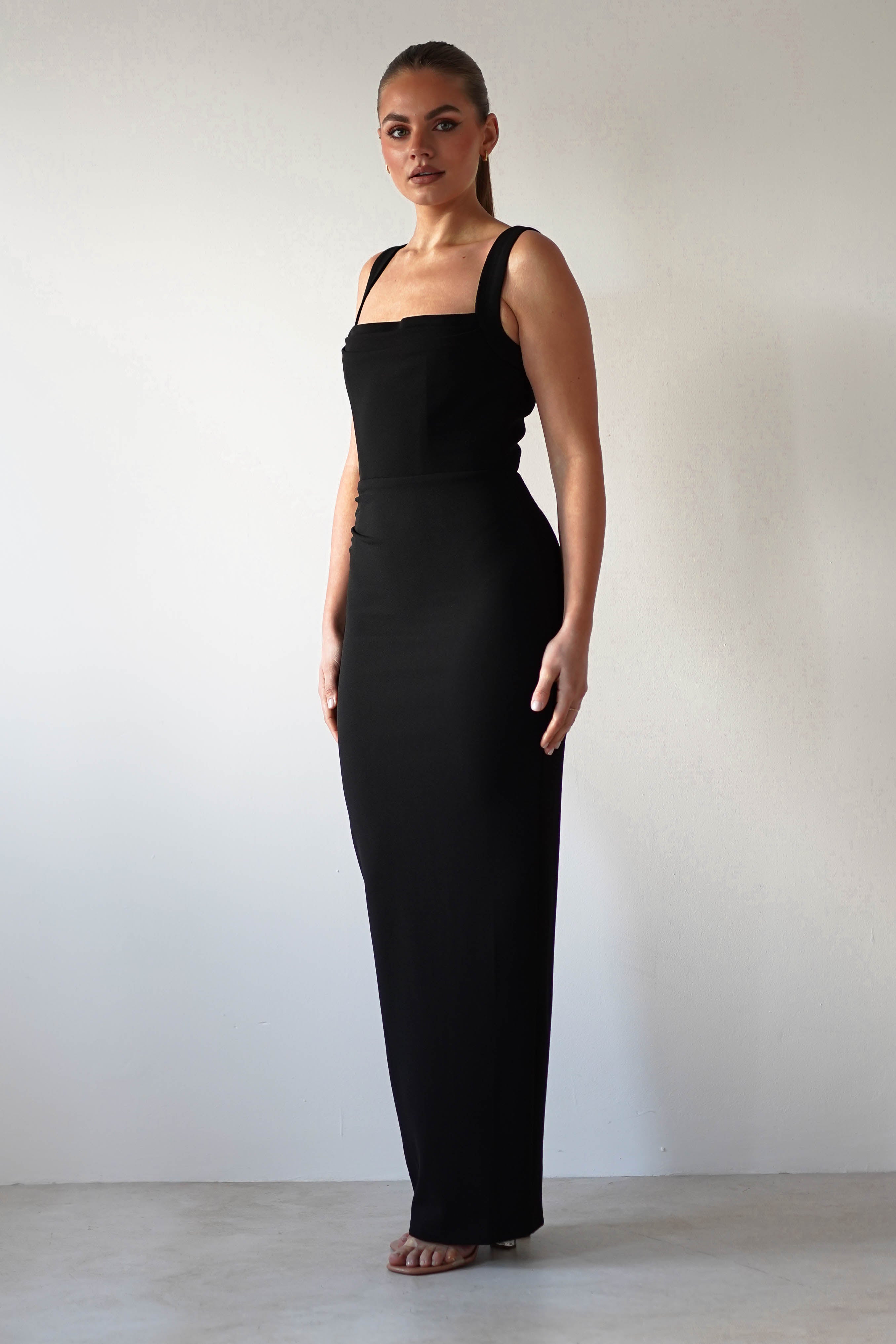 A person stands against a plain white background wearing a glamorous, long black dress with wide straps and a bodycon fit. They look directly at the camera with a neutral expression, their hair styled back neatly.