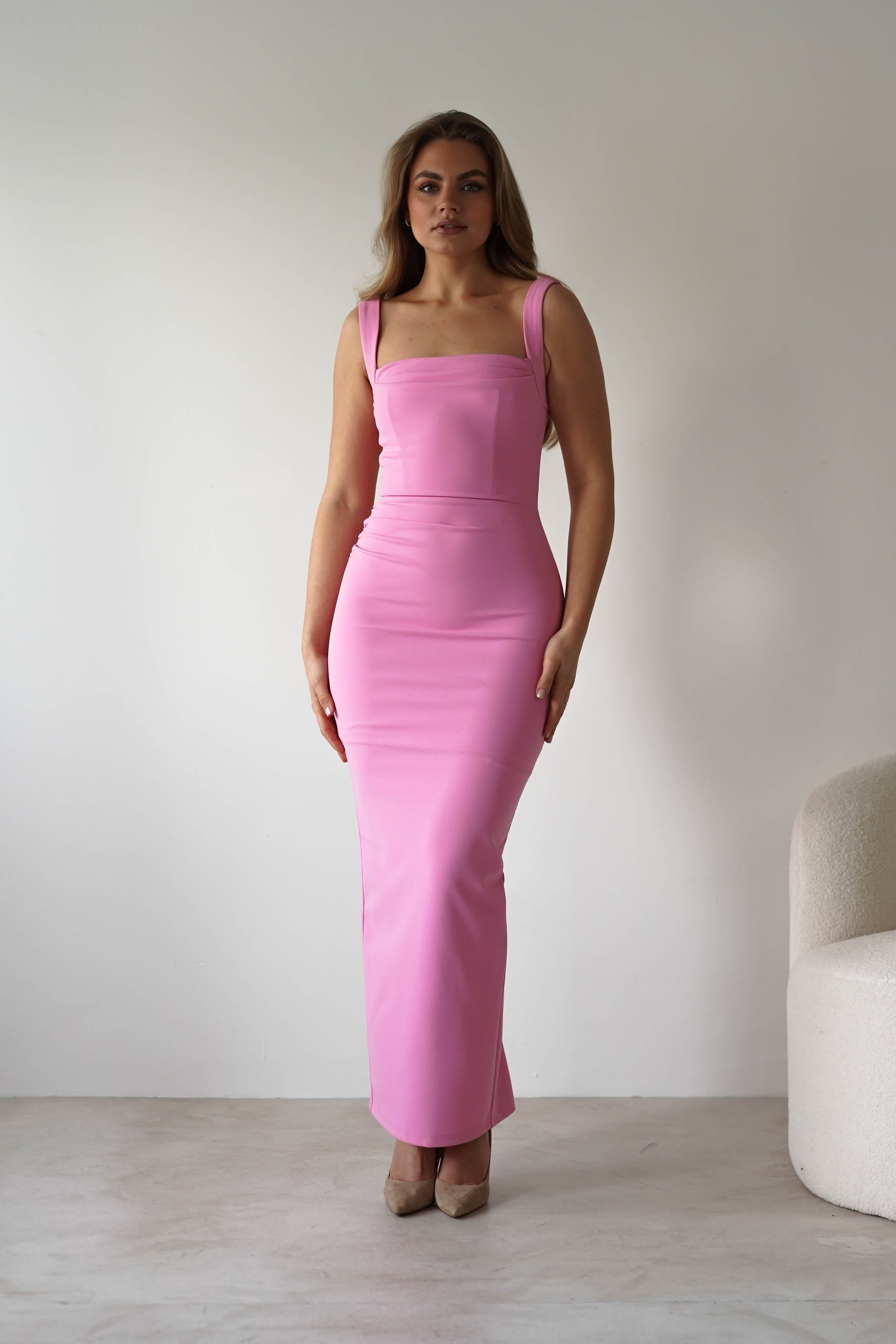 A woman stands in a minimalist room wearing a fitted, sleeveless, floor-length pink square neck dress and beige high heels. She faces forward with relaxed arms, making this the perfect elegant look for a wedding guest. A light-colored chair is visible on the right.
