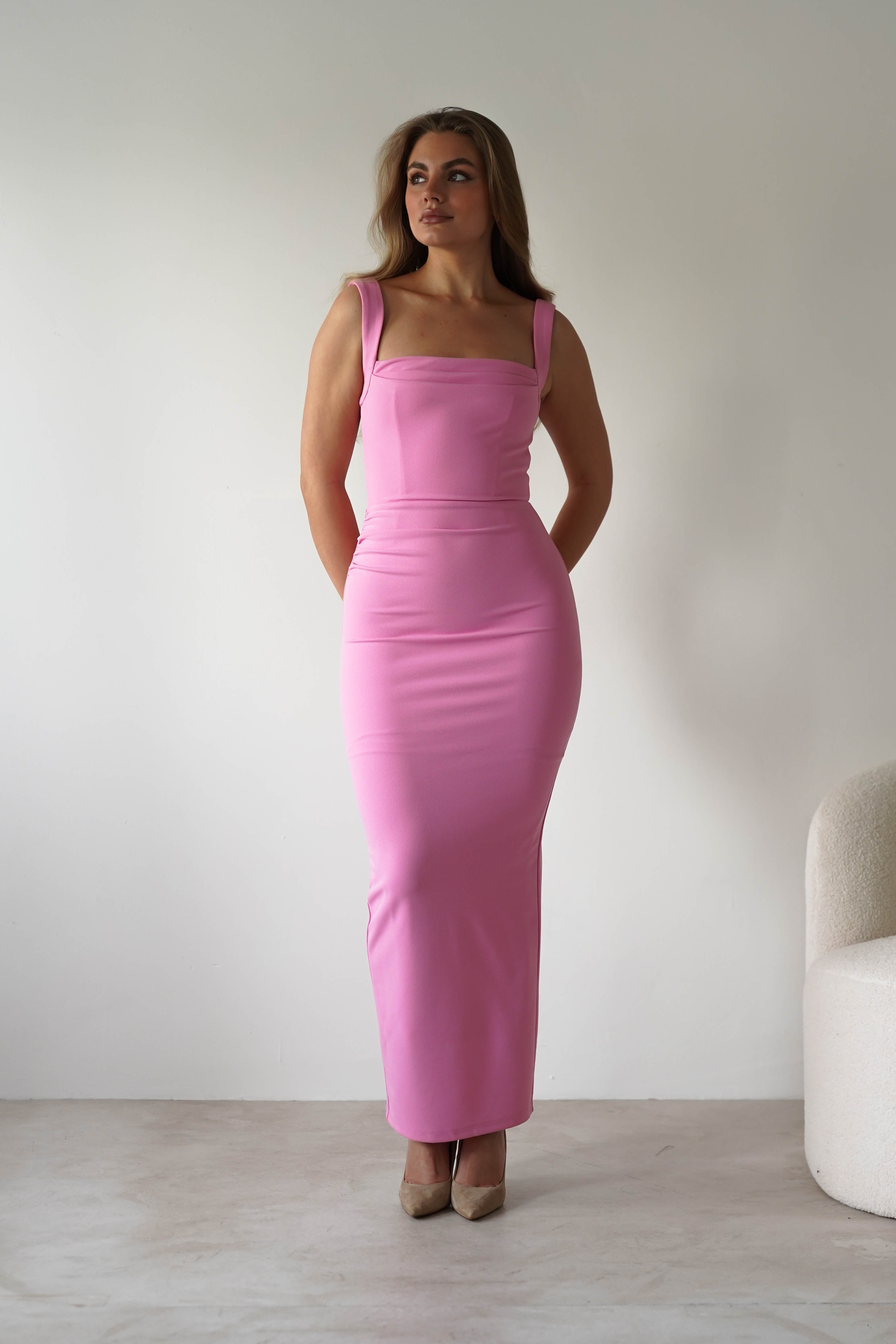 A woman wearing a fitted, sleeveless pink square neck dress stands against a plain white background, hands behind her back and looking to the side. A light-colored chair is partially visible beside her—perfect for a stylish wedding guest look.