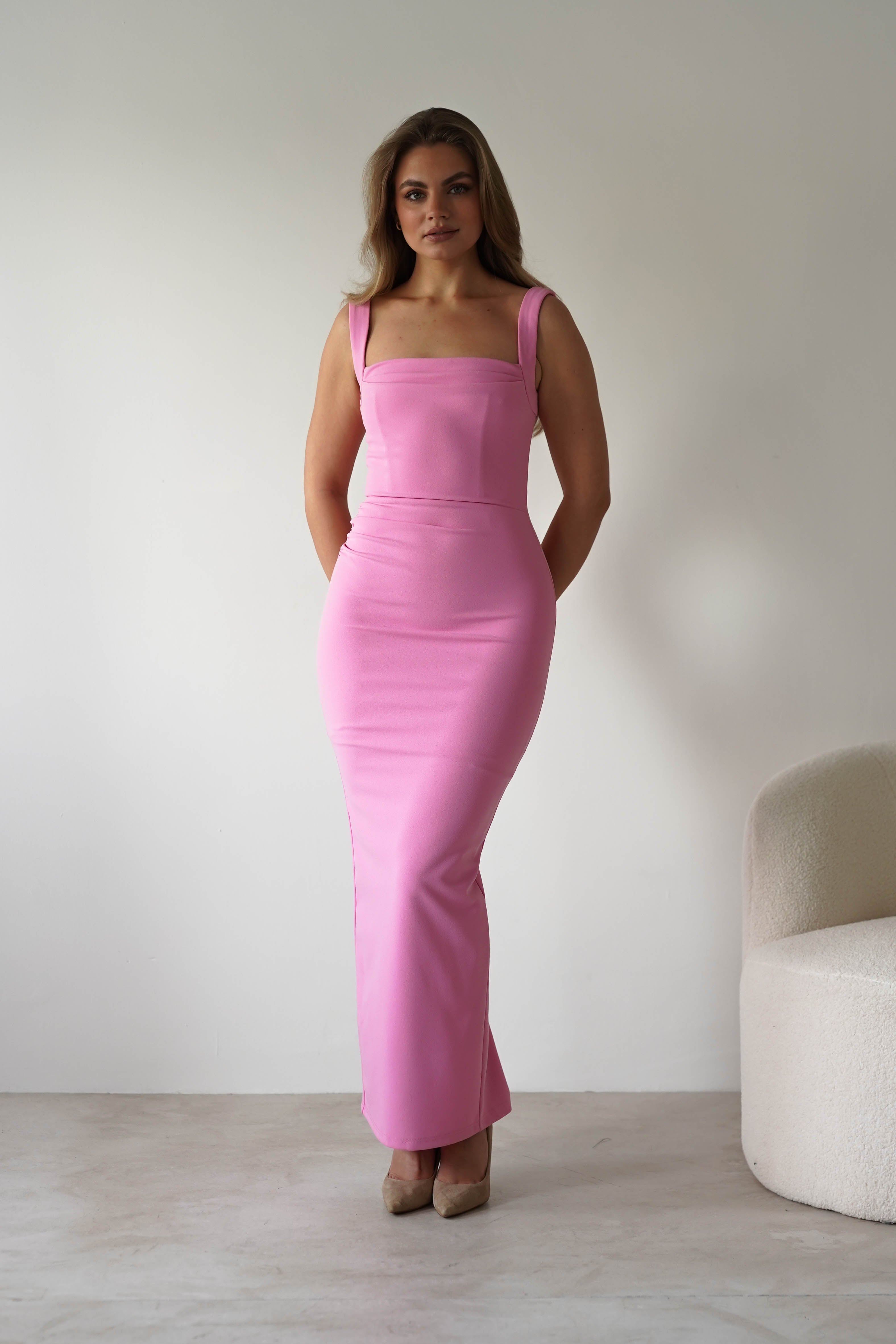 A woman stands indoors wearing a fitted, sleeveless pink square neck dress and beige heels. She poses with her hands behind her back against a plain white background, with a white chair to the right—a chic look for a wedding guest.