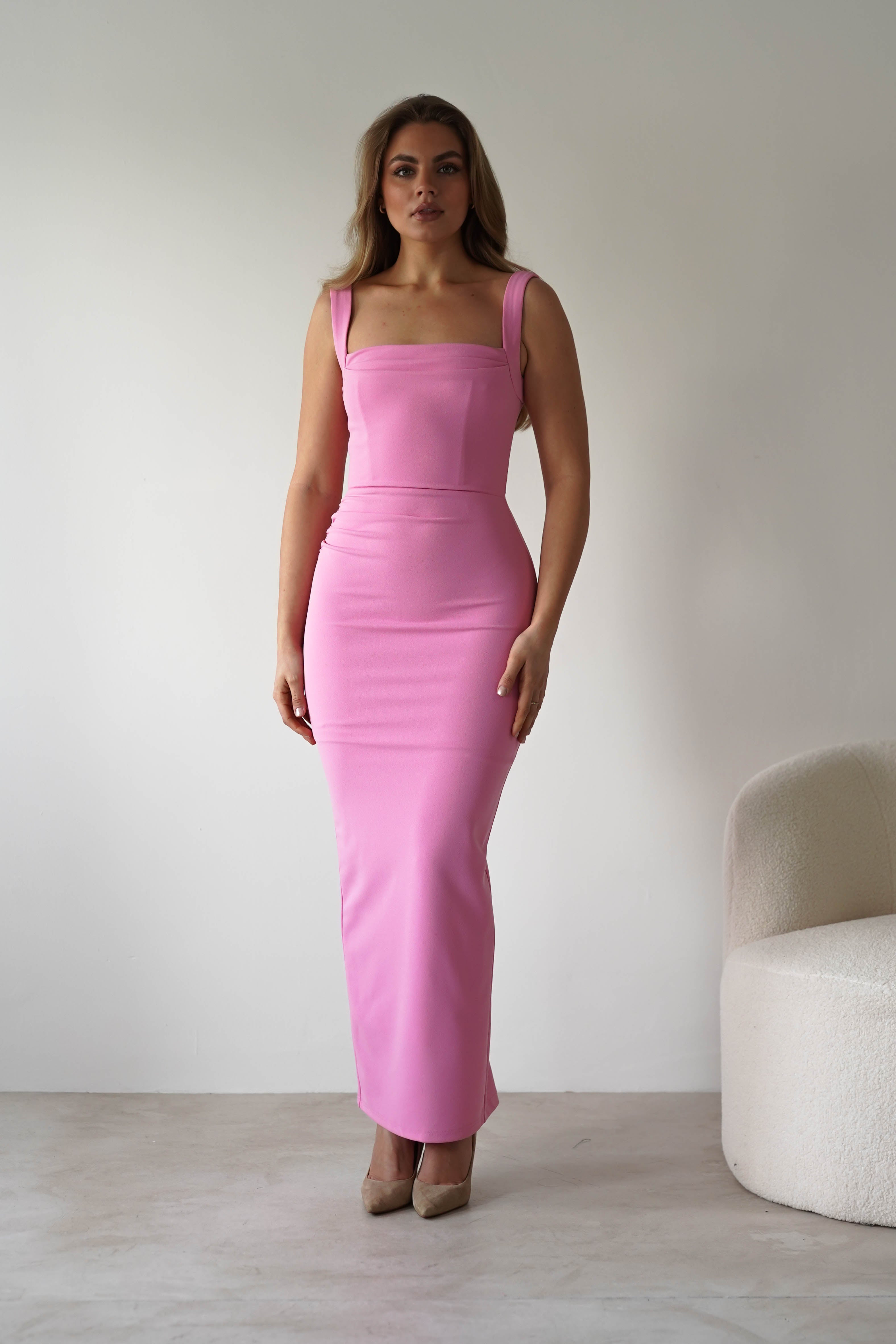 A woman stands indoors near a white curved chair, wearing a pink square neck dress with a bodycon fit that reaches her ankles—an elegant choice for a wedding guest. Her long, wavy hair cascades against a plain white wall.