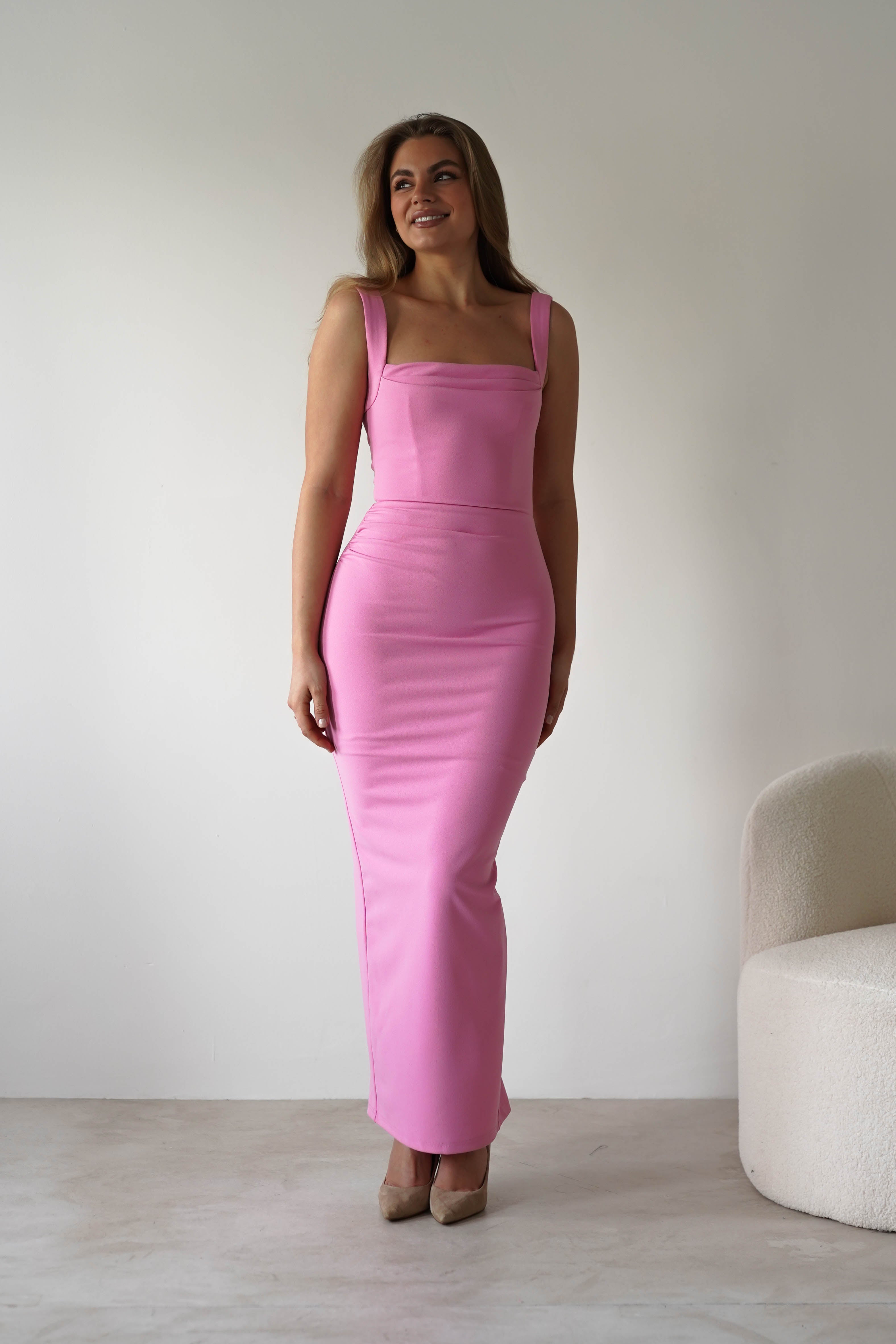 A woman stands indoors in a form-fitting, sleeveless, floor-length pink square neck dress and beige heels, smiling slightly. She is positioned near a white textured chair against a plain, light-colored background—an elegant choice for a wedding guest.