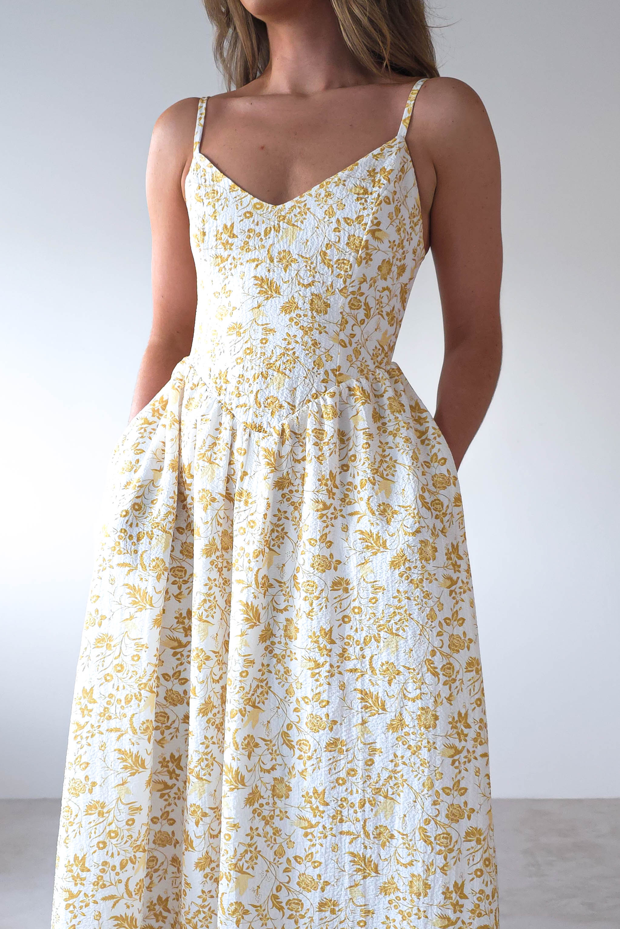 A woman in a white summer print maxi dress with yellow floral patterns features adjustable ties and pockets, standing against a plain background. The dress has a fitted bodice that flares into an A-line skirt. Her hair is loose and wavy, embodying an effortless style perfect for wedding guests.