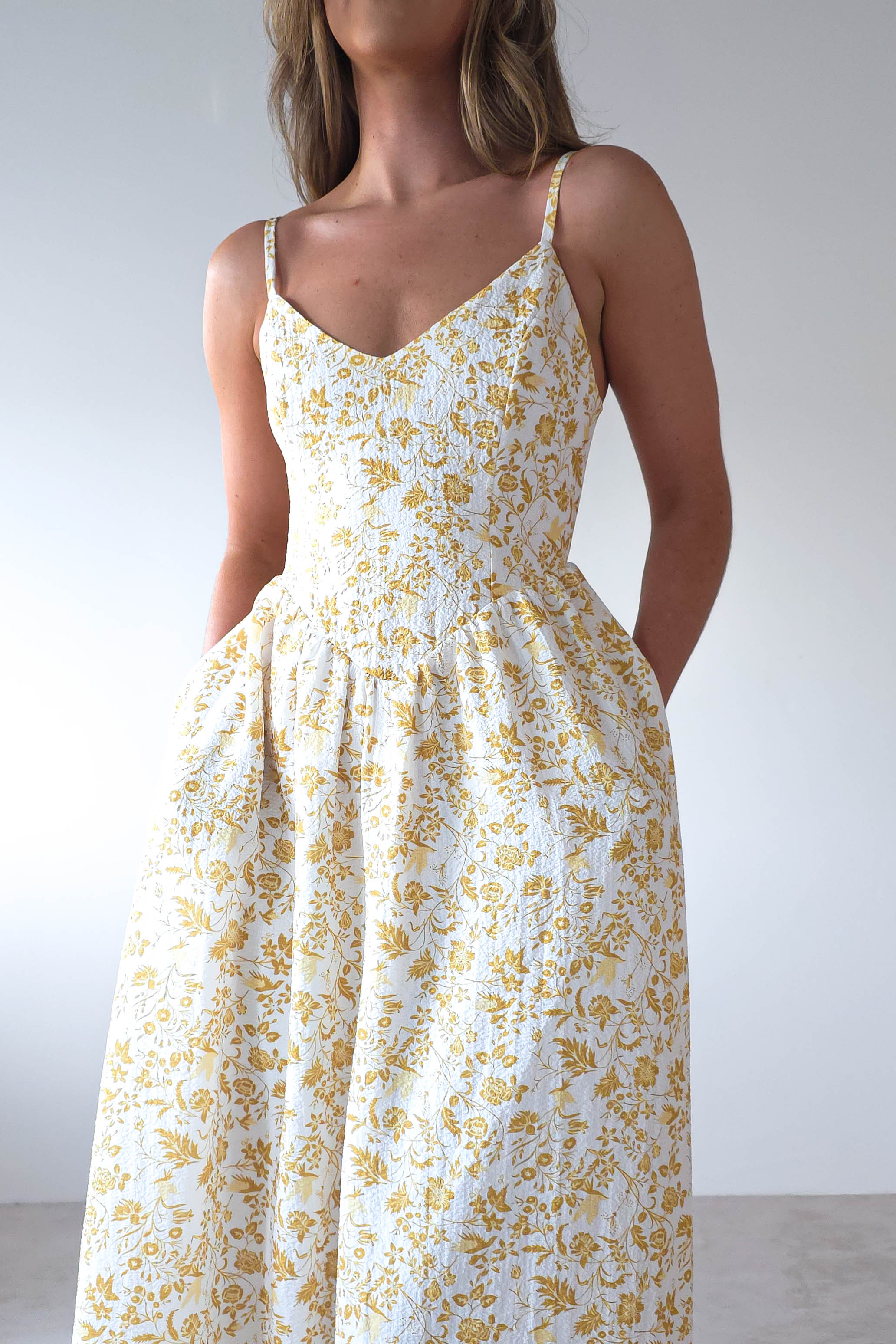 A woman wearing a sleeveless, white summer print maxi dress with a yellow floral pattern poses against a plain, light-colored background. The dress features adjustable ties, thin straps, a fitted bodice, and a flared skirt with pockets—perfect for wedding guests.